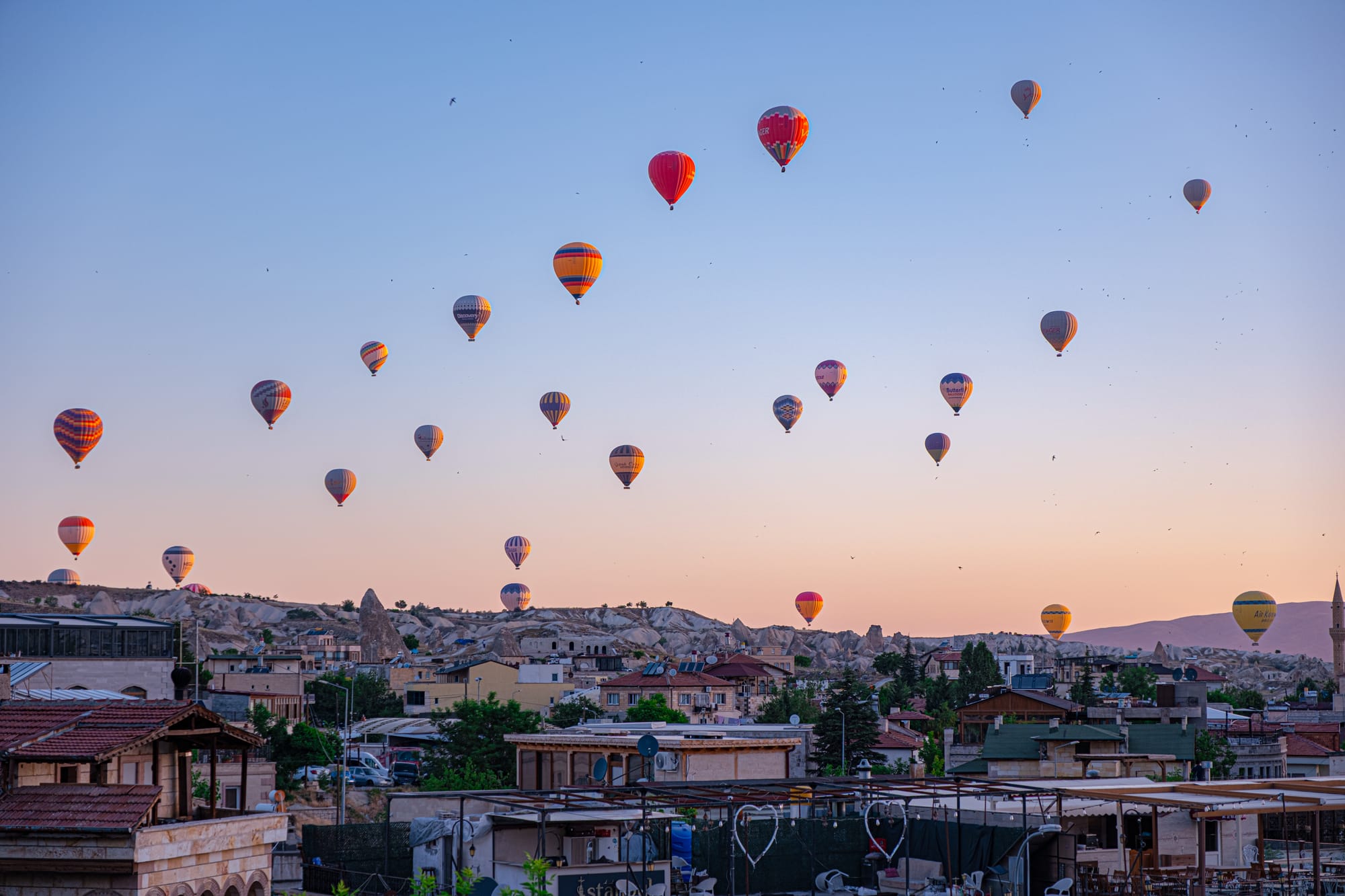 Morning view over Göreme, Cappadocia: dozens of colorful hot air balloons float across the sky at sunrise. Below, rooftops and houses sit against the backdrop of pale stone hills and ridges, the scene bathed in soft golden light