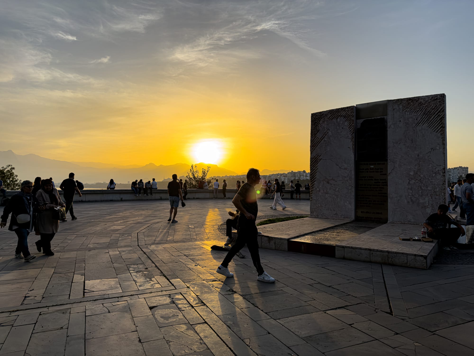 People strolling and sitting near a stone monument in Karaalioğlu Park, Antalya, silhouetted against a vivid orange sunset over the mountains and sea