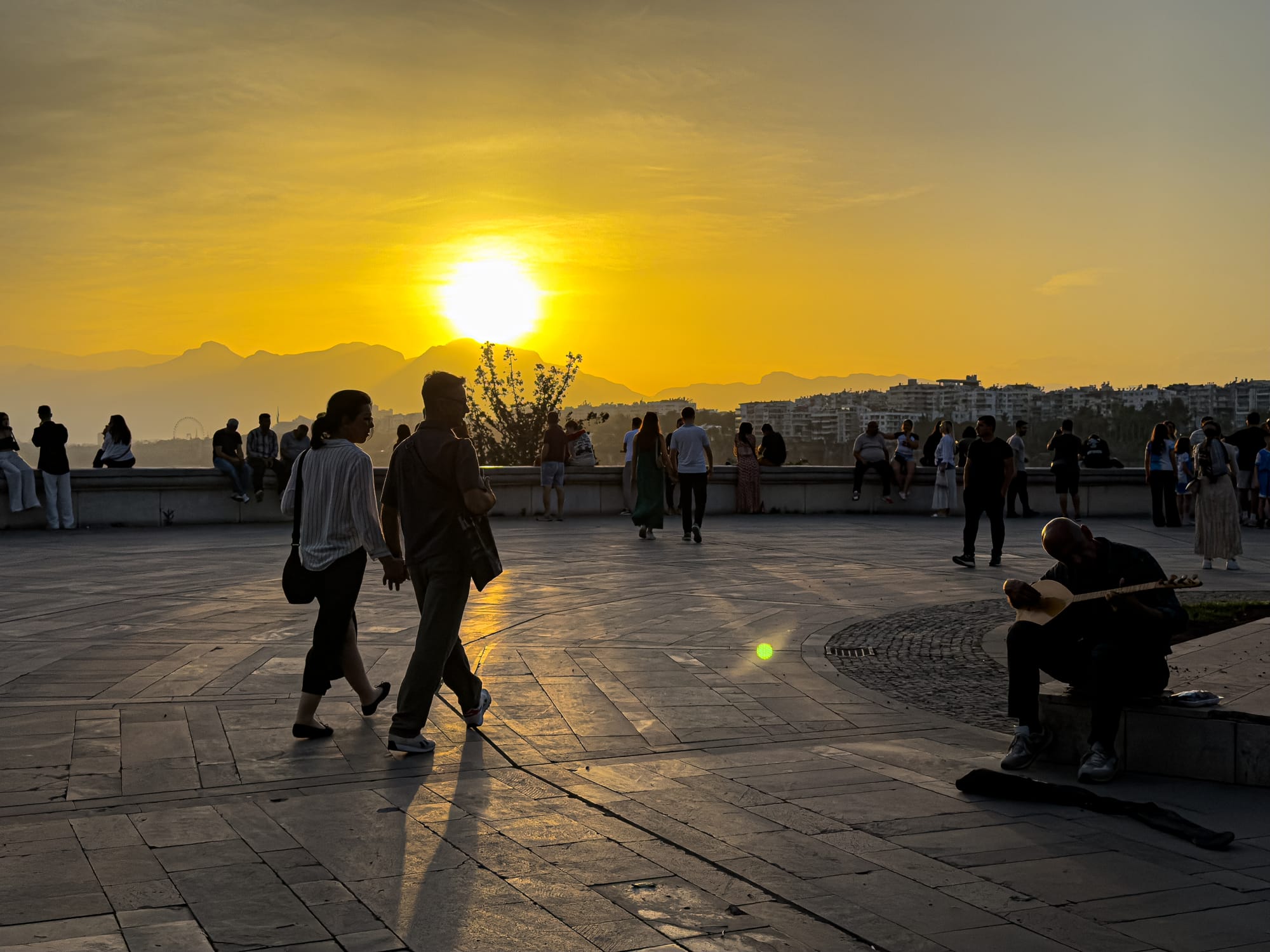 People gather at Karaalioğlu Park in Antalya during sunset, with golden light casting long shadows and a street musician playing a traditional instrument