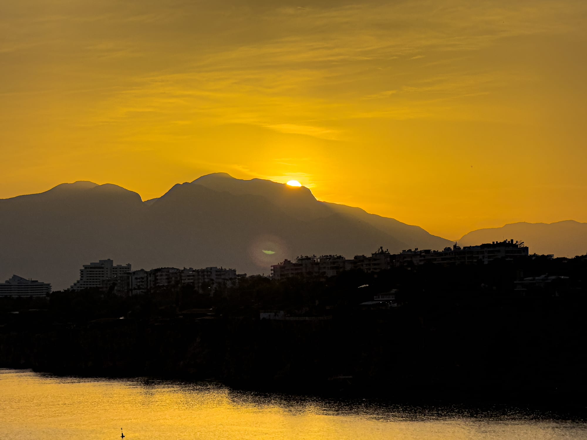 Golden sunset over Antalya’s mountainous horizon, with the city’s silhouette in the foreground and golden light reflecting on the water