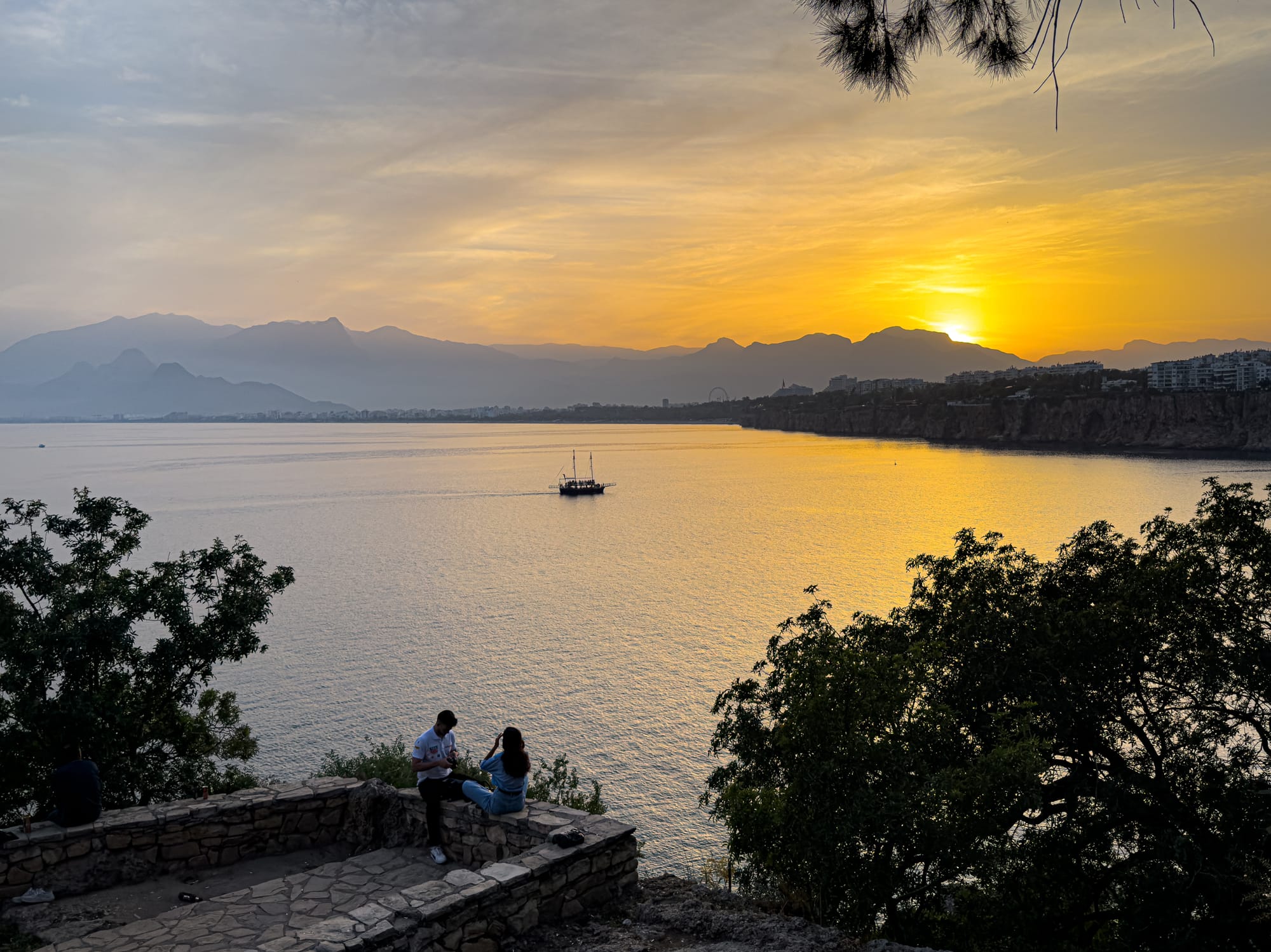 Golden hour view from Karaalioğlu Park in Antalya, with people sitting on a stone terrace, a sailboat on the sea, and the sun setting behind distant mountains