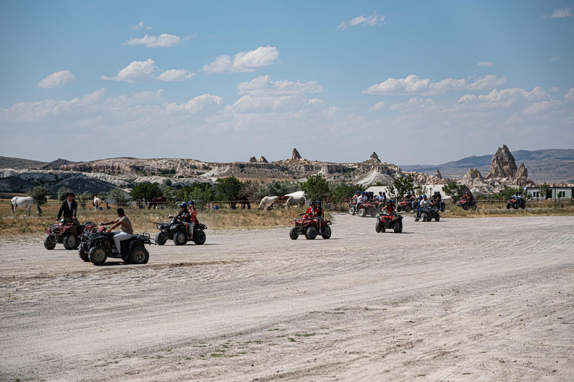 Large group of ATV riders driving across a dusty plain in Cappadocia with rock formations and grazing horses in the distance