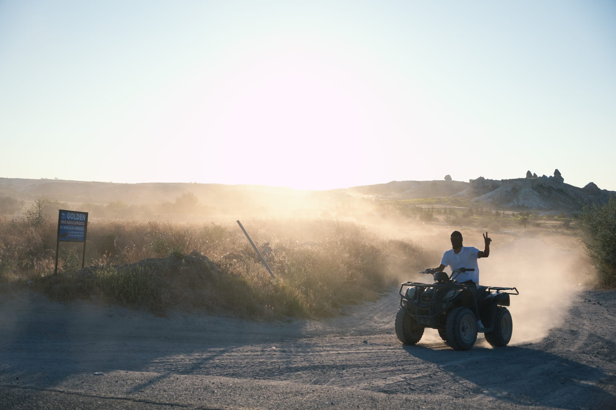 ATV rider on a dirt path in Cappadocia at sunset, raising a hand in a peace sign as dust rises in the golden light