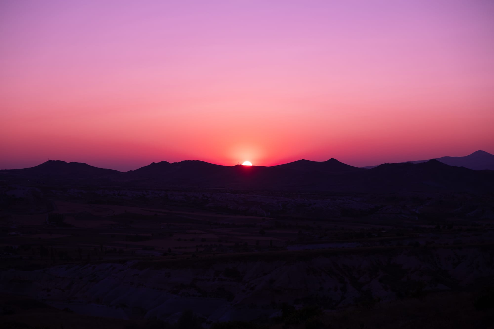 Sunset over Cappadocia with the sun setting behind dark mountain ridges and the sky glowing in pink and purple tones