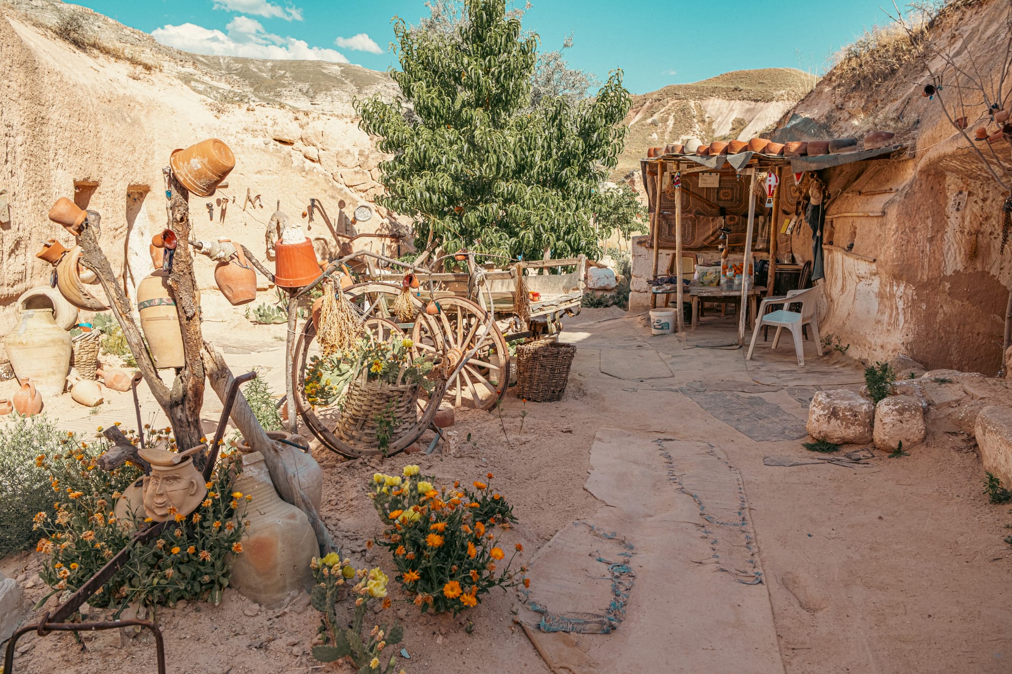 Courtyard of Tarihi Ev Historical House in Çavuşin, Cappadocia, Turkey, featuring a wooden cart, terracotta pots, woven baskets of flowers, hanging rustic tools, and a shaded seating area set against stone walls and a backdrop of rolling hills