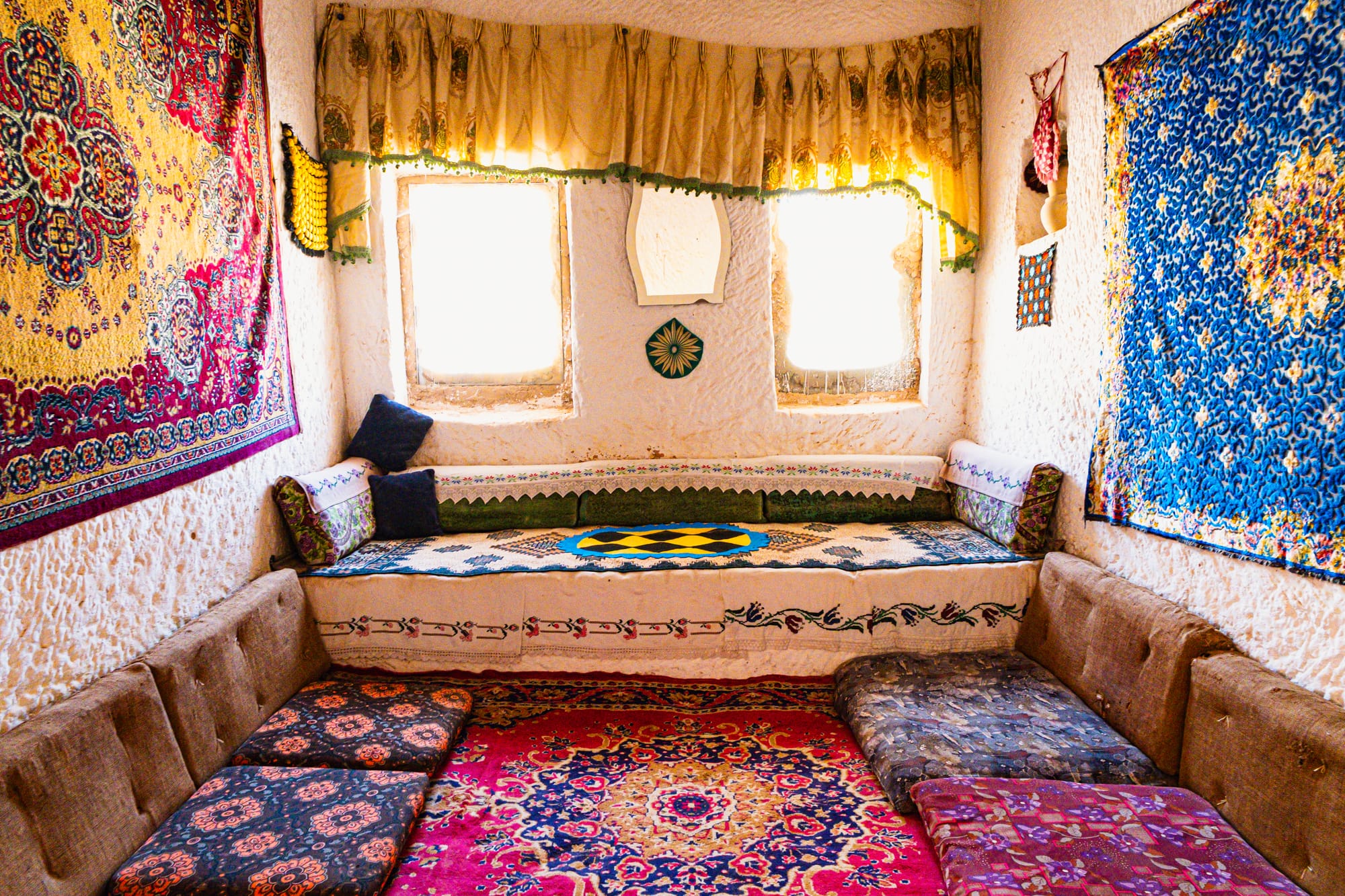 Cozy interior room at Tarihi Ev Historical House in Çavuşin, Cappadocia, featuring low cushioned seating, vibrant patterned rugs, and colorful wall tapestries illuminated by soft light from two windows