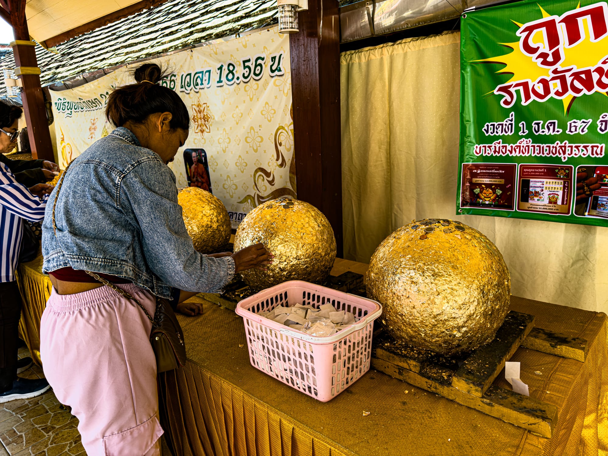A woman applies gold leaf to large golden sacred stones displayed on a table at a Chiang Mai temple, with a basket of gold leaf sheets beside her