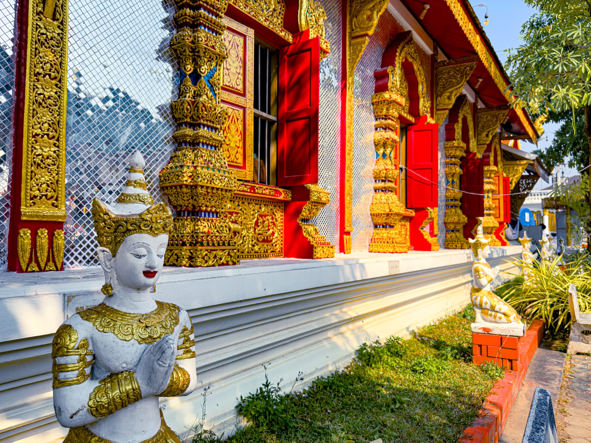 A richly decorated Chiang Mai temple with bright red shutters, golden ornate carvings, and mirrored mosaic walls, accompanied by white guardian statues with gold accents in prayer positions along the exterior walkway