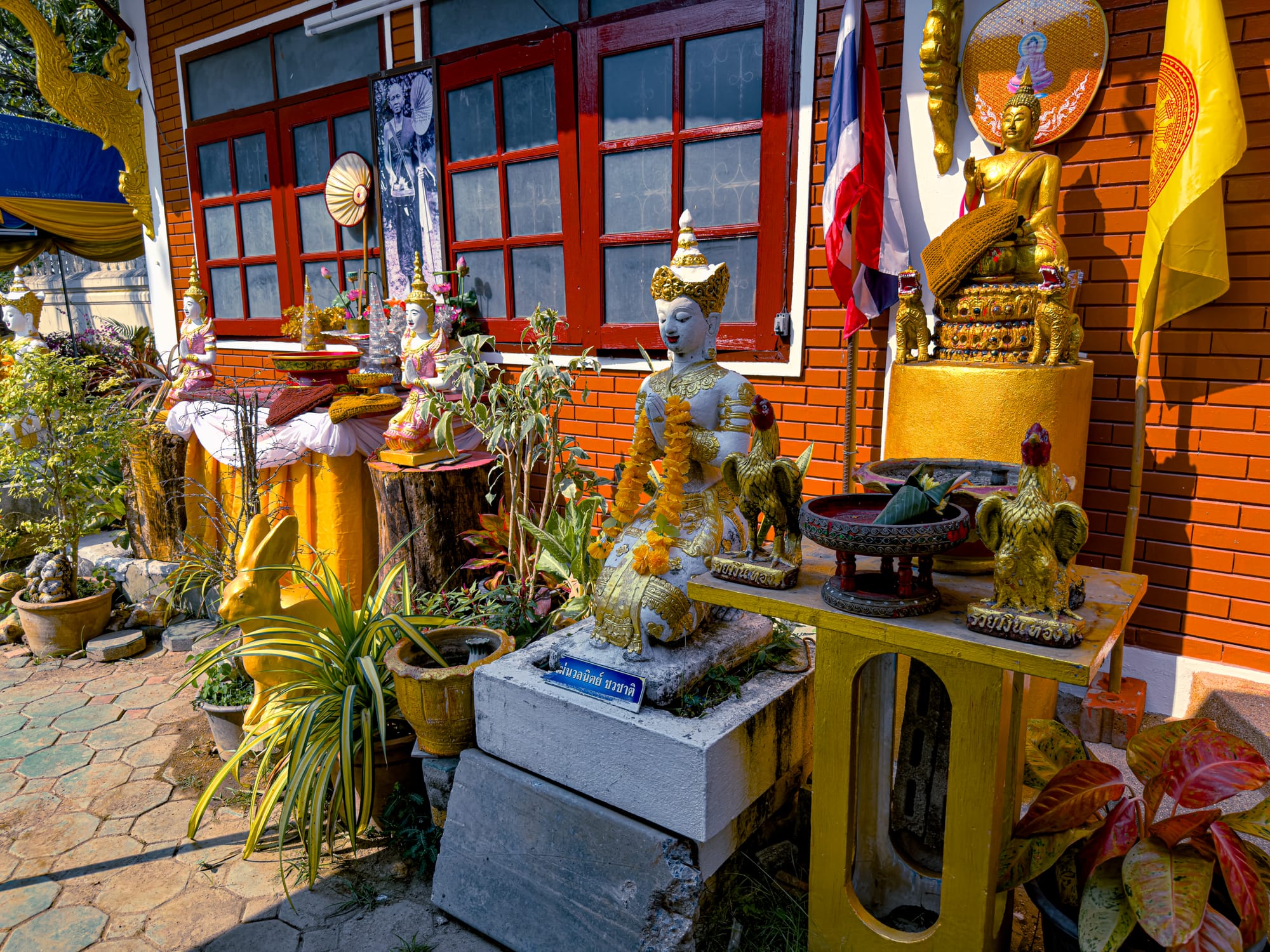 Outdoor temple shrine in Chiang Mai featuring golden Buddha statues, white guardian figures with floral garlands, rooster figurines, and offerings set against a red brick wall with flags