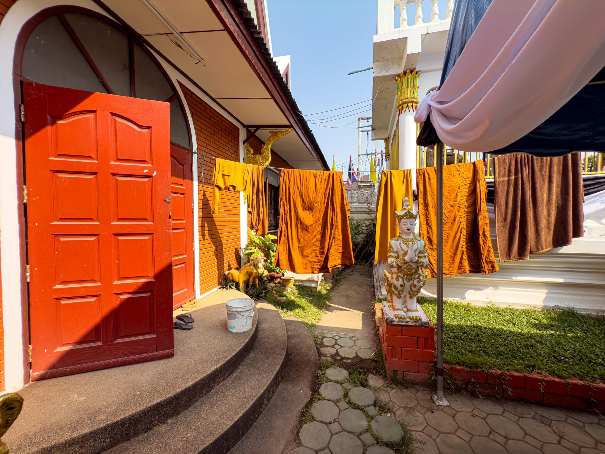 Bright orange and brown monk robes drying on clotheslines between temple buildings, with a red door, golden statue, and small walkway in view