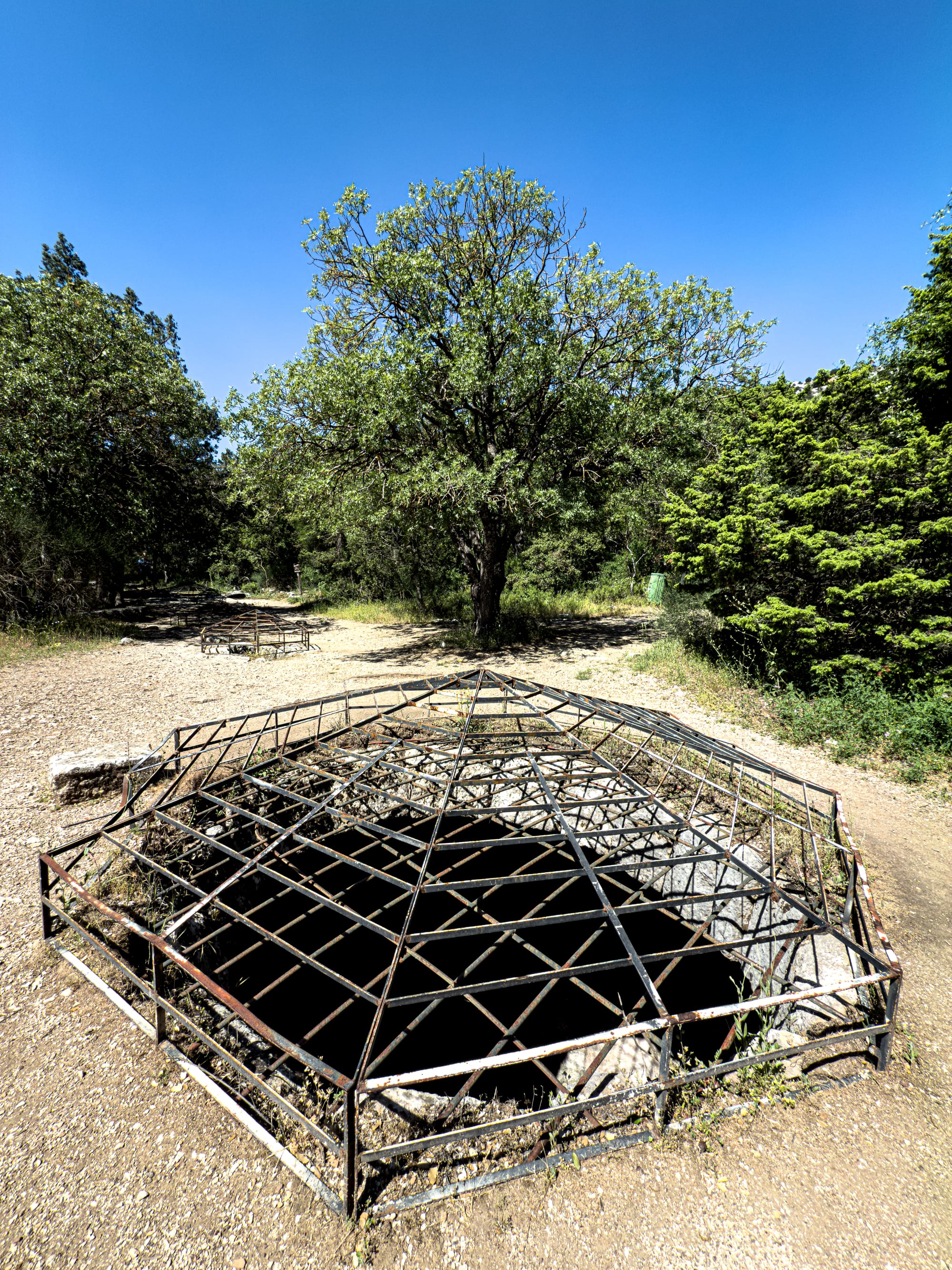 Stone-lined ancient cistern with a protective iron grate, part of Termessos’ original water storage system, surrounded by trees and sunlit ground