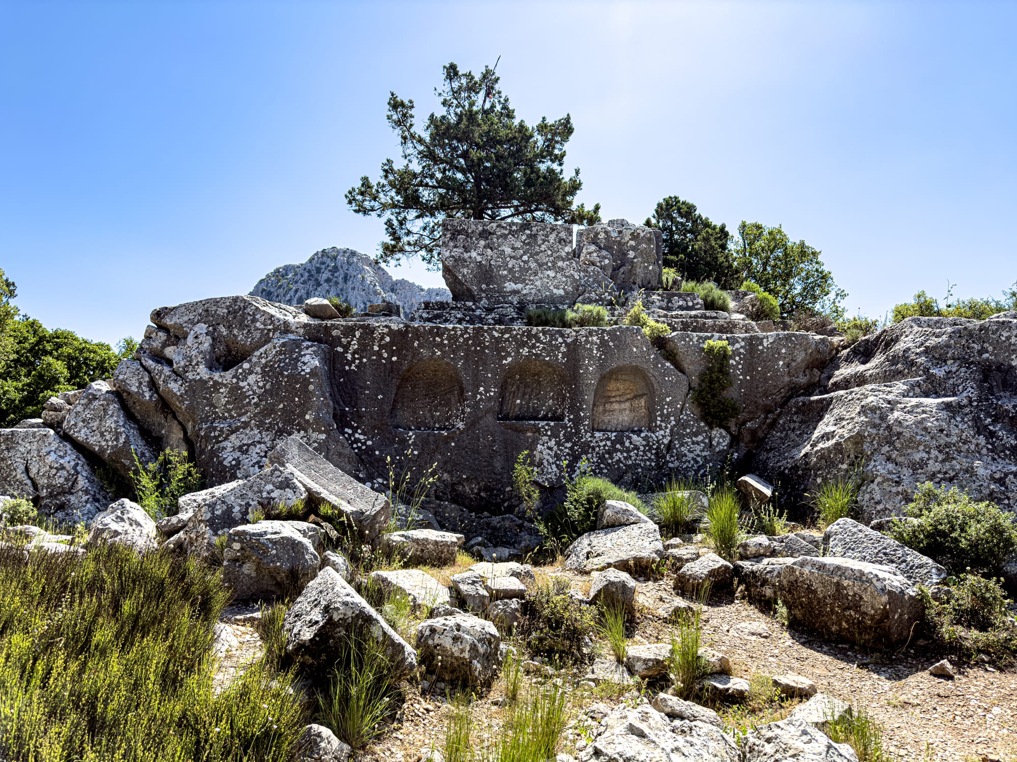 Rock-carved tombs at Termessos with arched niches cut directly into the limestone, surrounded by scattered boulders and wild grass under a bright blue sky