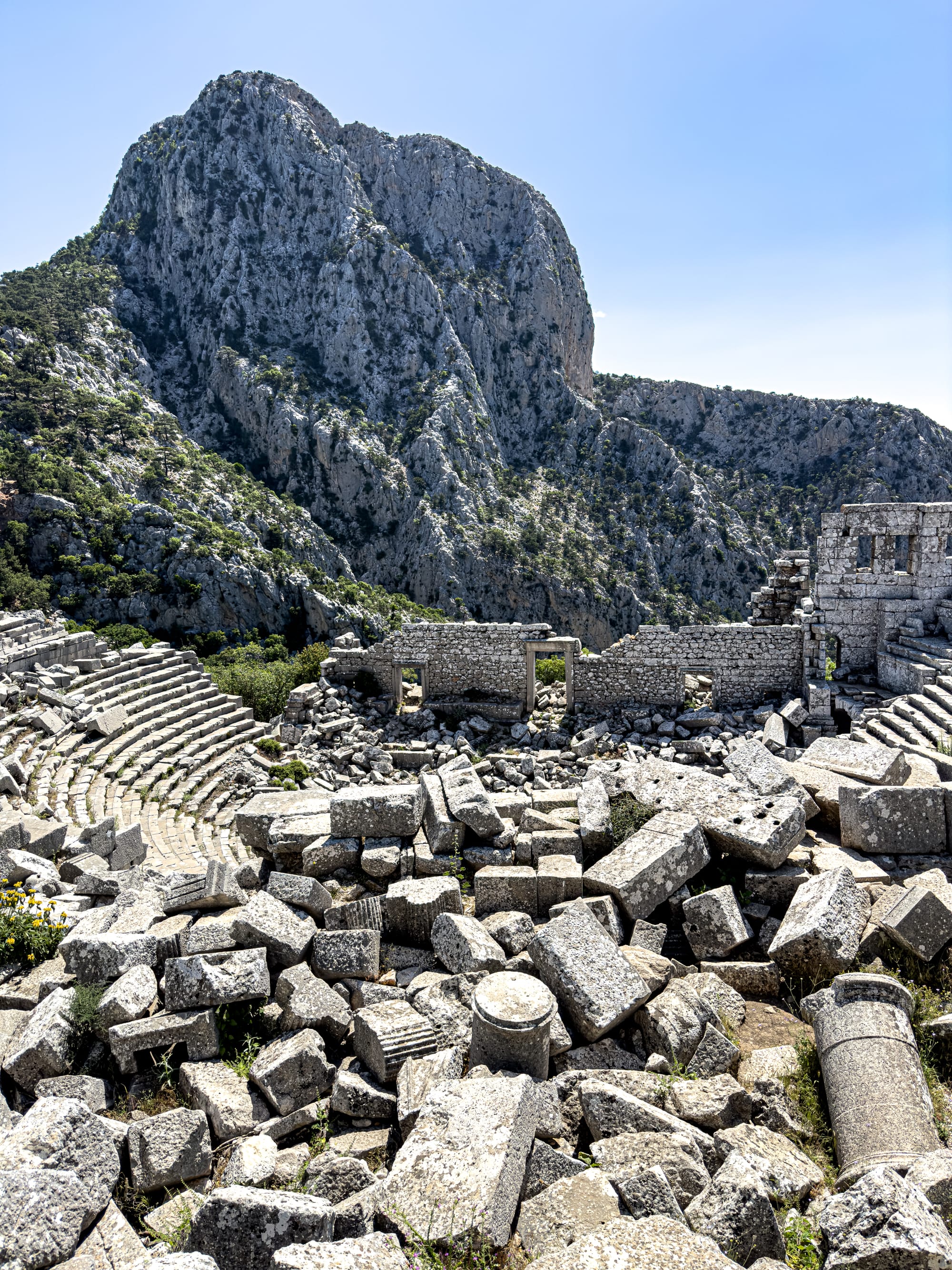 A wide view of the ancient amphitheater at Termessos with collapsed stone blocks in the foreground and a dramatic mountain backdrop in the distance