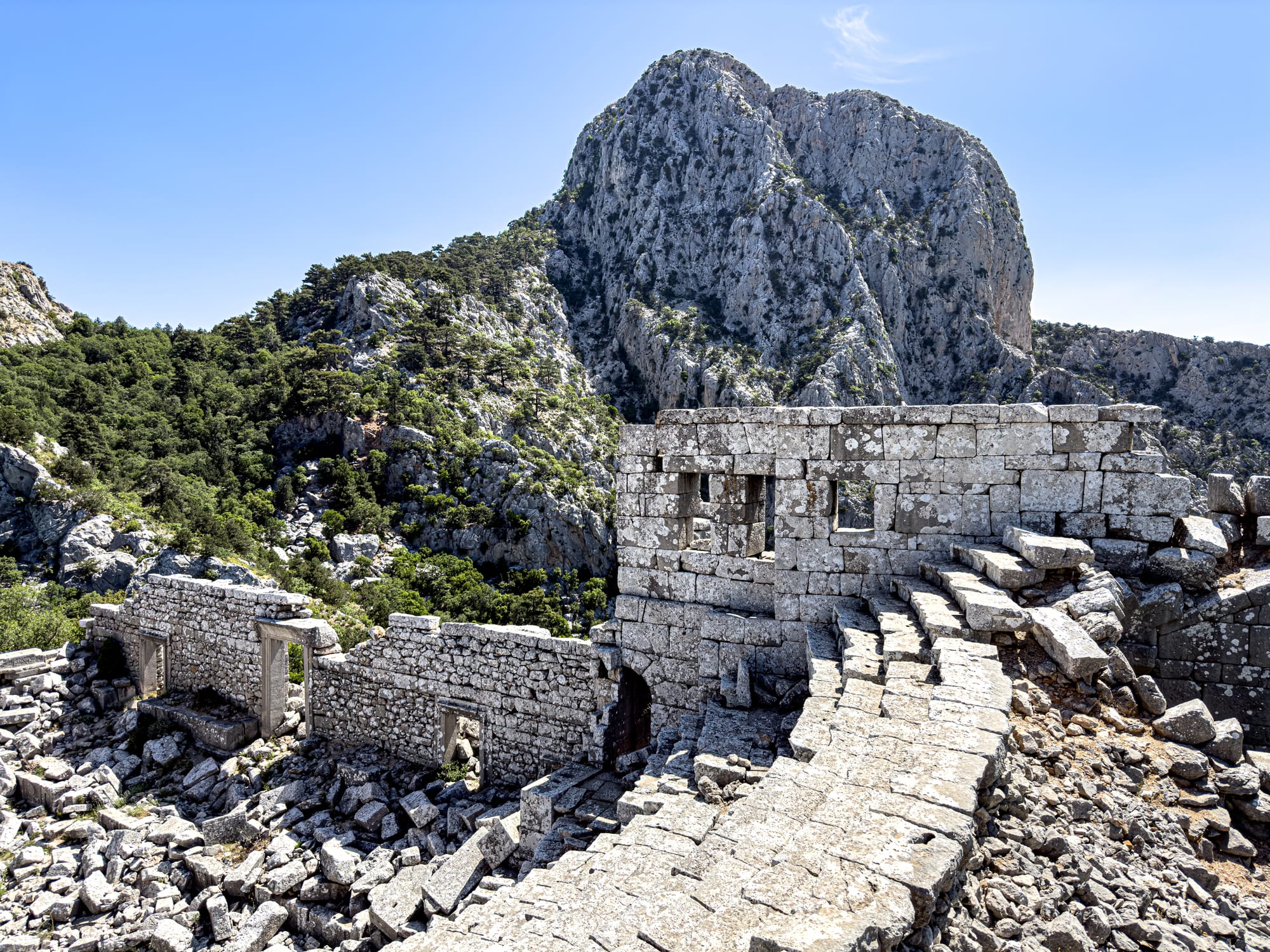 View of Termessos’ ancient stone structures perched along a mountainside, with remnants of stairways and doorways leading into thick green forest and dramatic limestone cliffs beyond