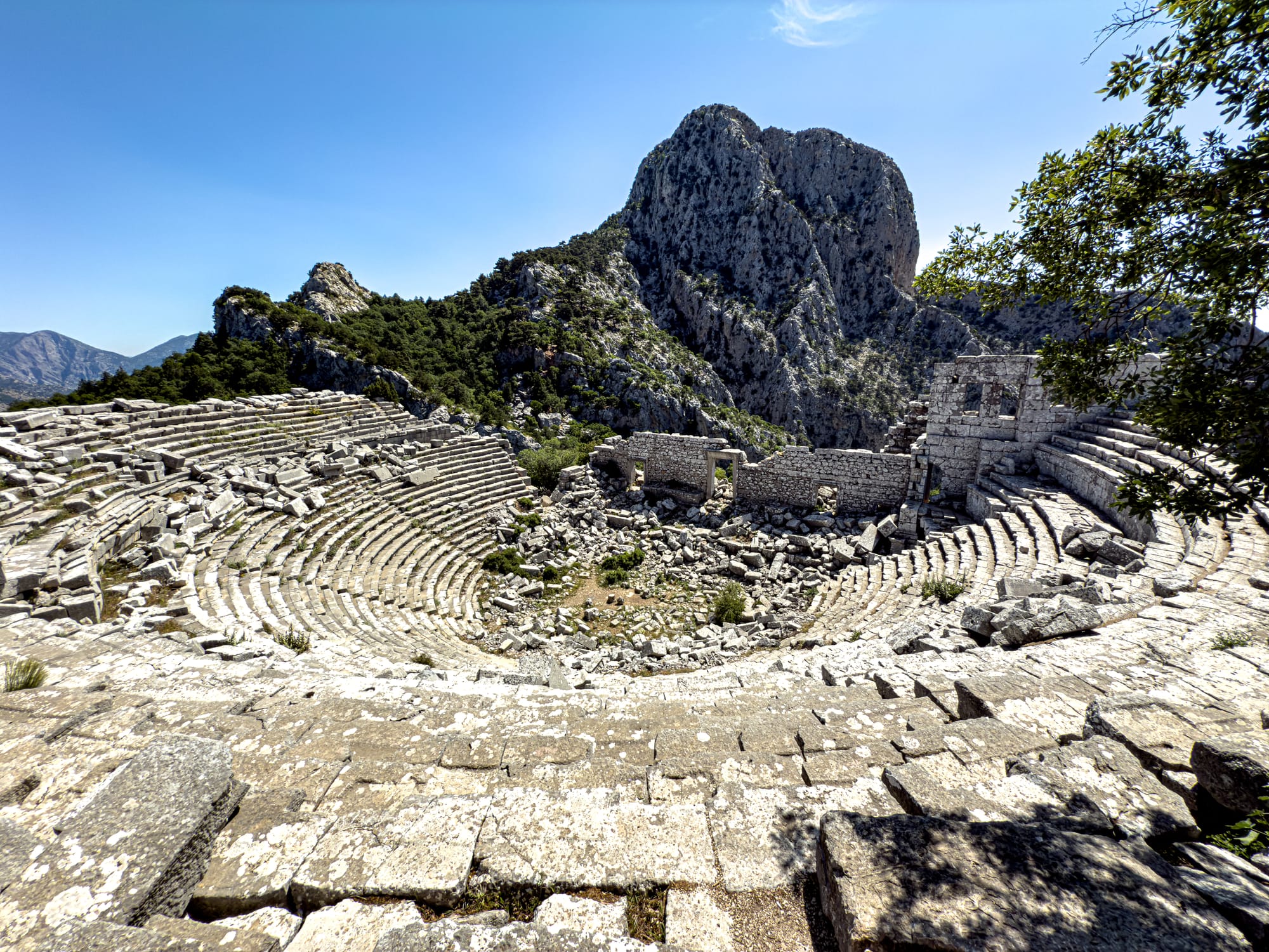 Wide-angle shot of the Termessos amphitheater with concentric stone seating, surrounded by pine-covered mountains and dramatic rocky cliffs