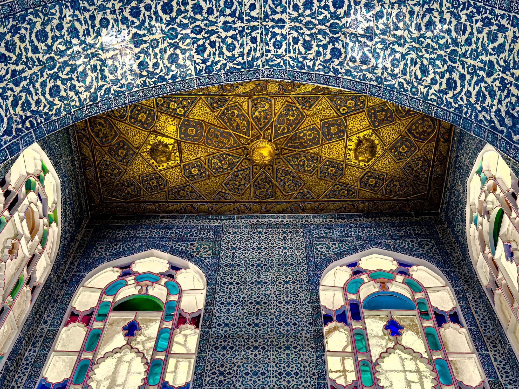 Ornate golden ceiling with geometric patterns and two stained glass windows framed by blue Iznik tiles at Topkapi Palace
