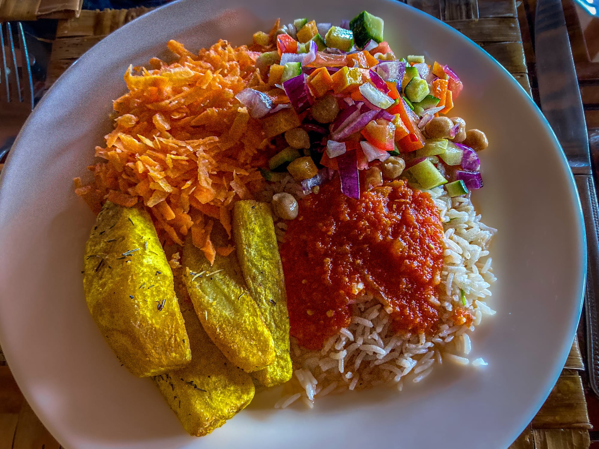 A white plate of food served at Voi Wildlife Lodge with basmati rice topped with tomato sauce, slices of golden fried potato, shredded carrots, and a salad of chopped cucumber, tomato, red onion, and chickpeas