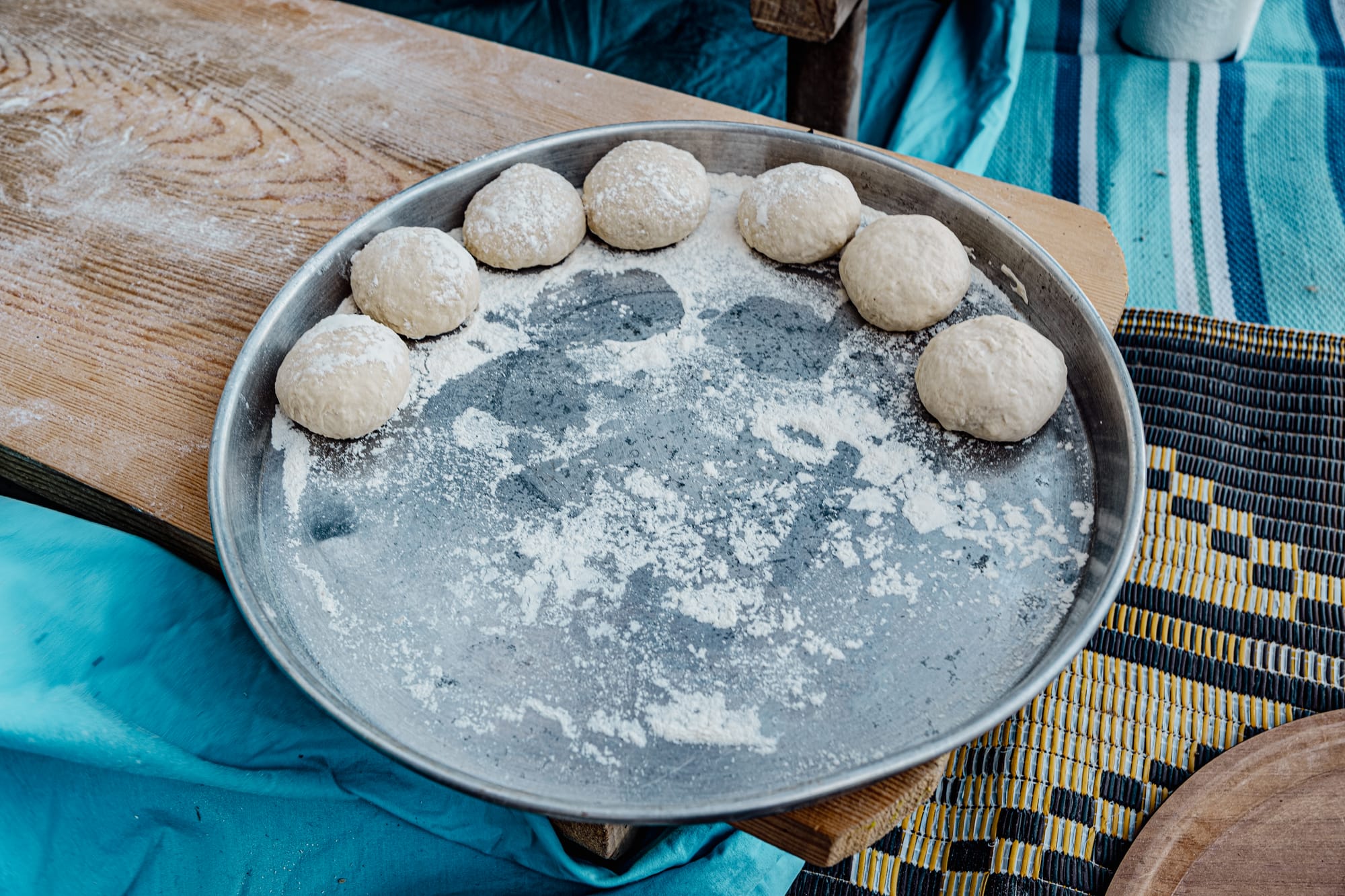 Close-up of a round metal tray holding several flour-dusted dough balls, placed on a wooden table with a woven mat and blue cloth underneath, prepared for making traditional Turkish gözleme
