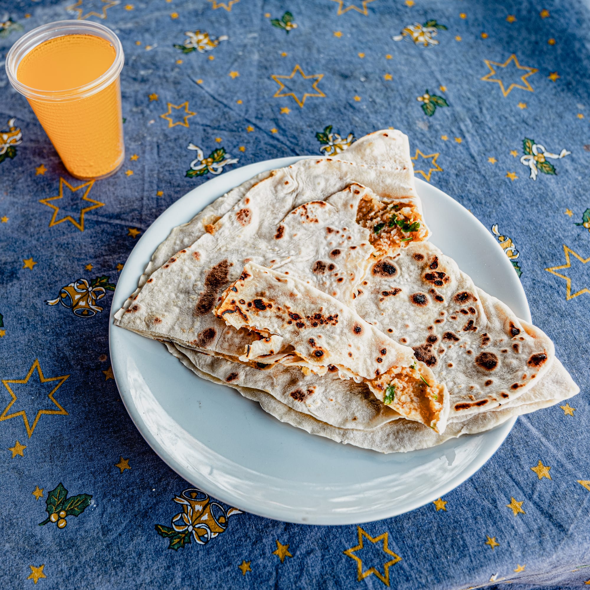 A plate of freshly cooked gözleme stuffed with herby potato filling, served alongside a cup of orange juice on a patterned blue tablecloth