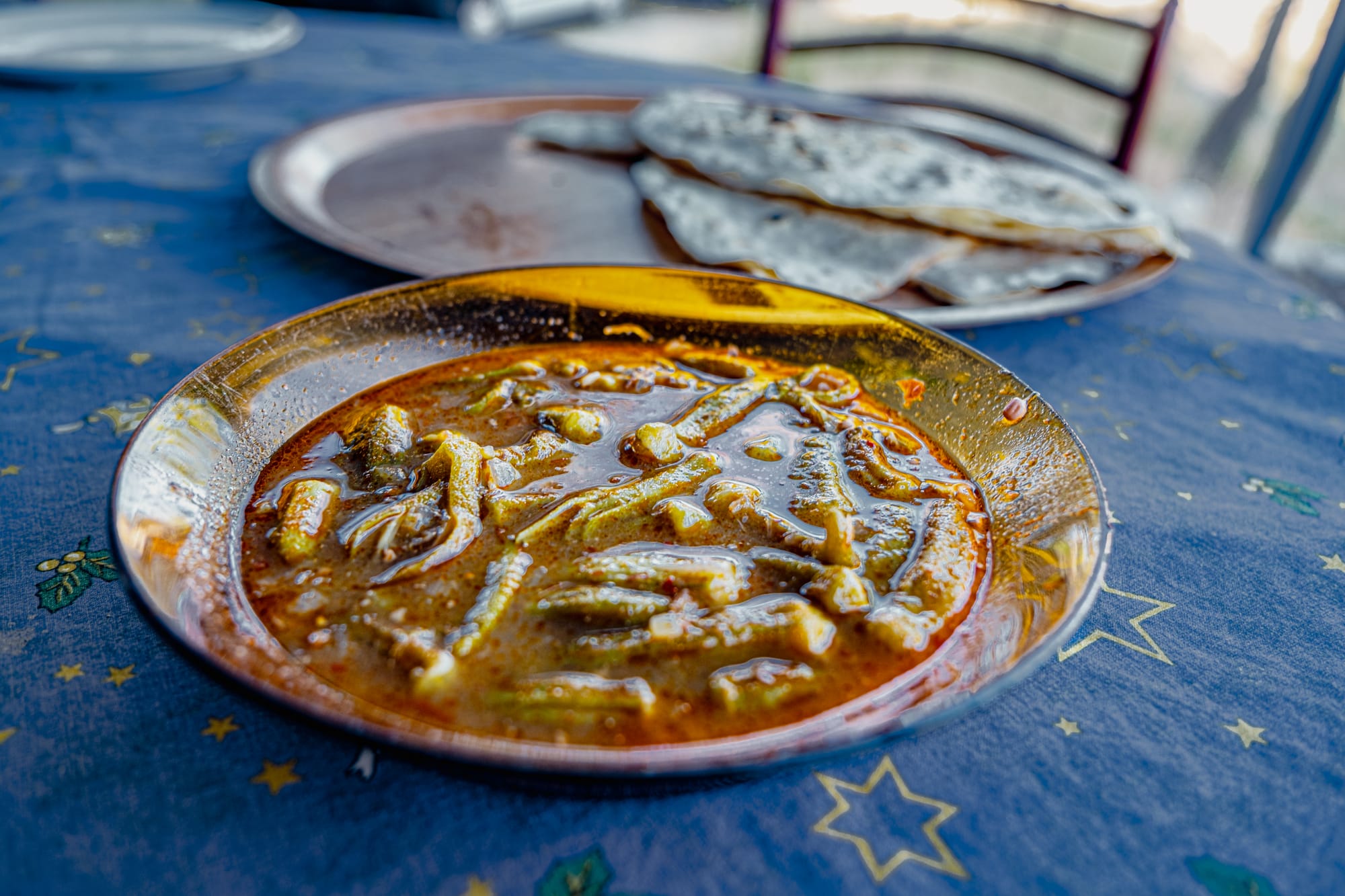 Close-up of a golden-brown okra stew in a shallow dish on a blue tablecloth, with folded gözleme visible in the background