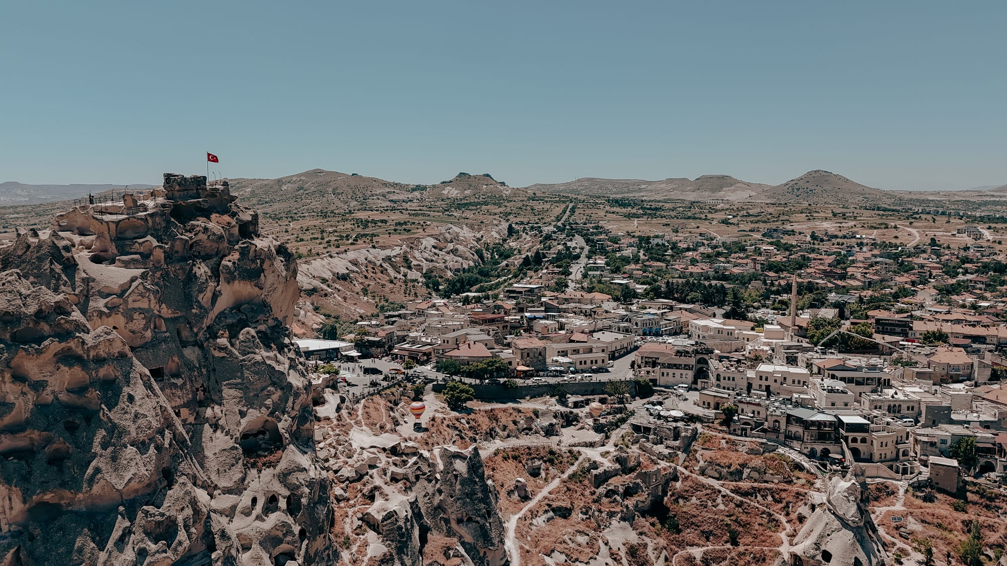 Uçhisar Castle Cappadocia aerial view showing summit with Turkish flag, village below, and connecting walking trails into valleys