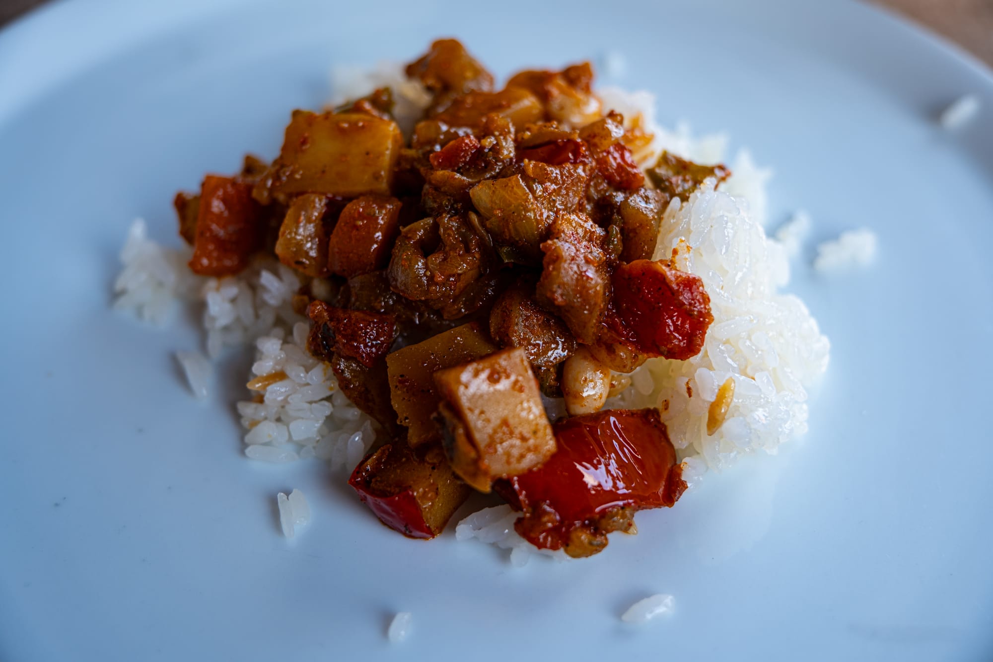 Close-up of a portion of vegetable stew with carrots, peppers, and potatoes in a rich sauce, served on top of white rice on a plain white plate