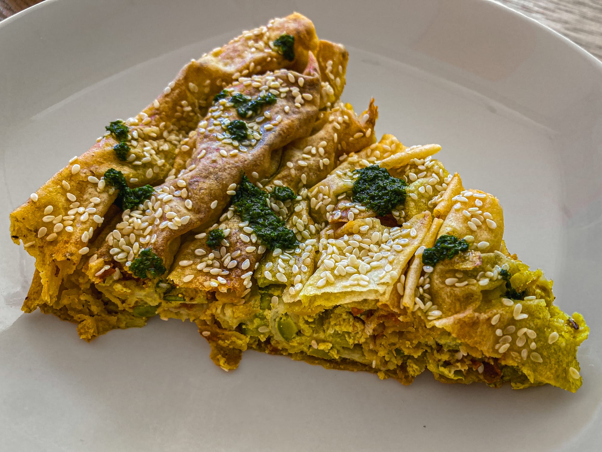 Slice of vegan banitsa topped with sesame seeds and drizzled with green pesto, filled with chickpea, zucchini, and sundried tomato