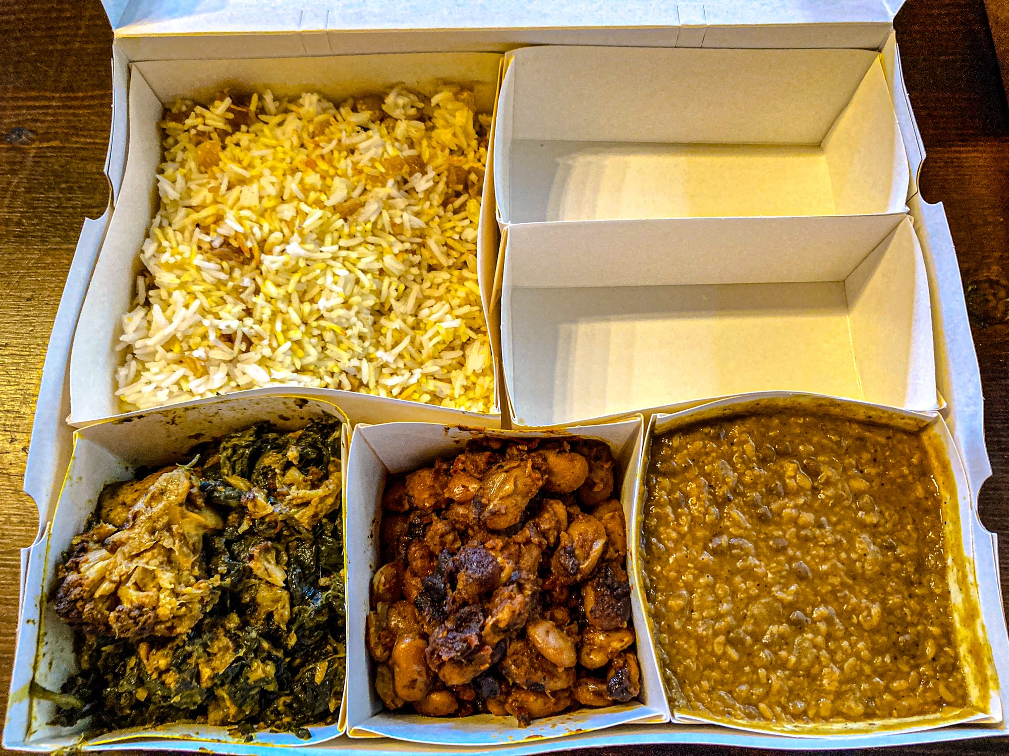 Boxed takeaway meal from Avalon Coliving’s curry night in Bansko, featuring compartments of yellow spiced rice, a lentil dal, a bean curry, and a vegetable curry with greens