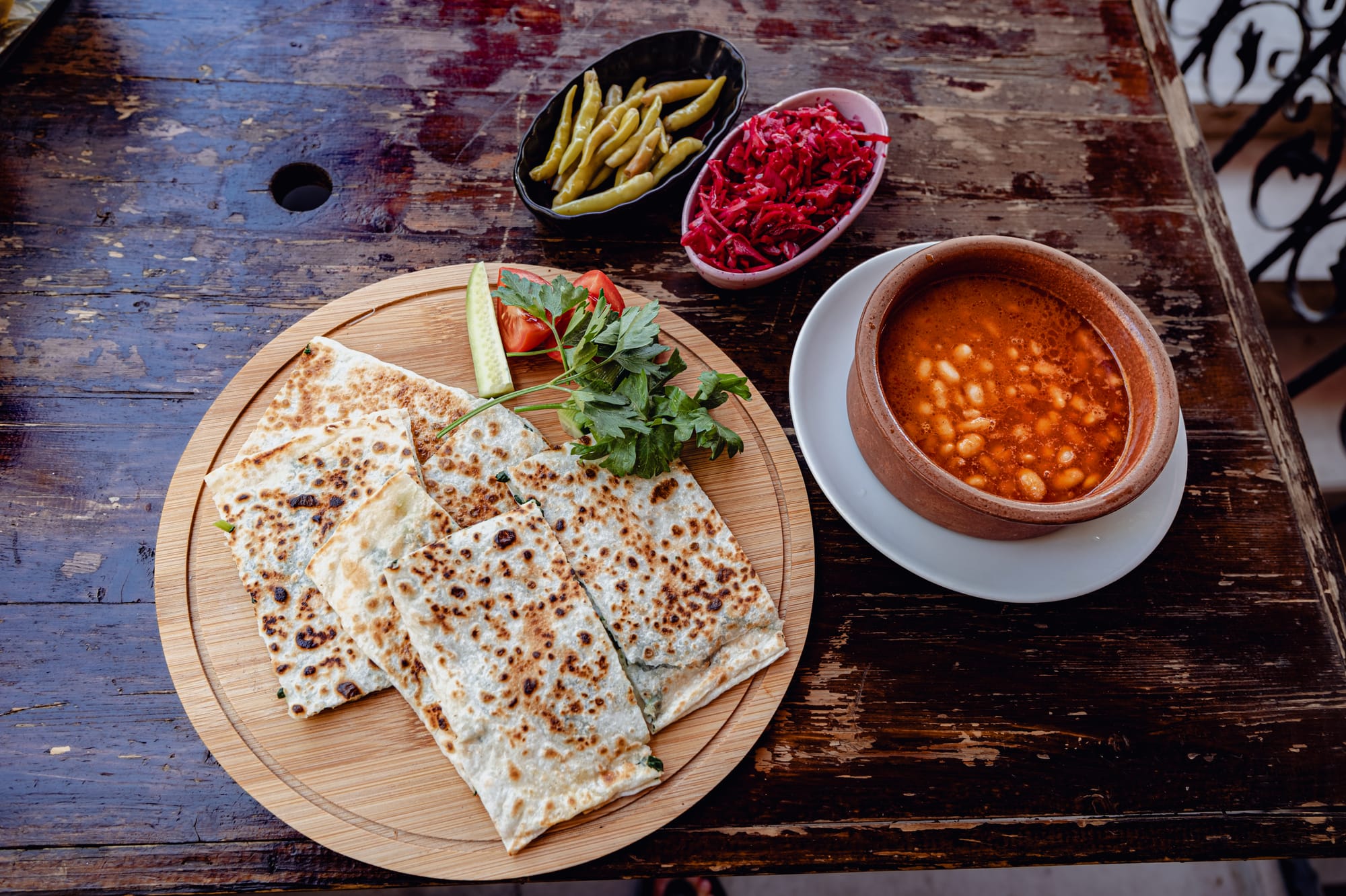 Plate of vegan gözleme, a Turkish flatbread stuffed with herby potato filling, garnished with parsley, tomato, and cucumber, served with a bowl of tomato-based bean stew, homemade pickles, and shredded red cabbage on a rustic wooden table at Neman Cave Restaurant in Avanos, Cappadocia