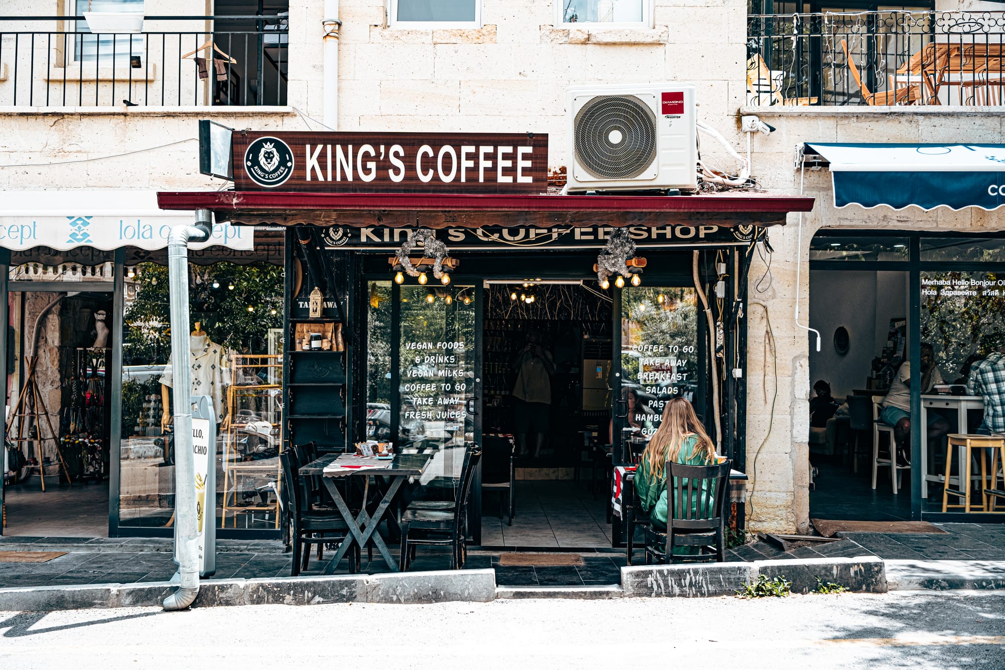 Street view of King’s Coffee Shop in Göreme, Cappadocia, with outdoor seating, a wooden sign reading “KING’S COFFEE,” and large windows listing menu highlights such as vegan foods, vegan milks, coffee to go, and fresh juices