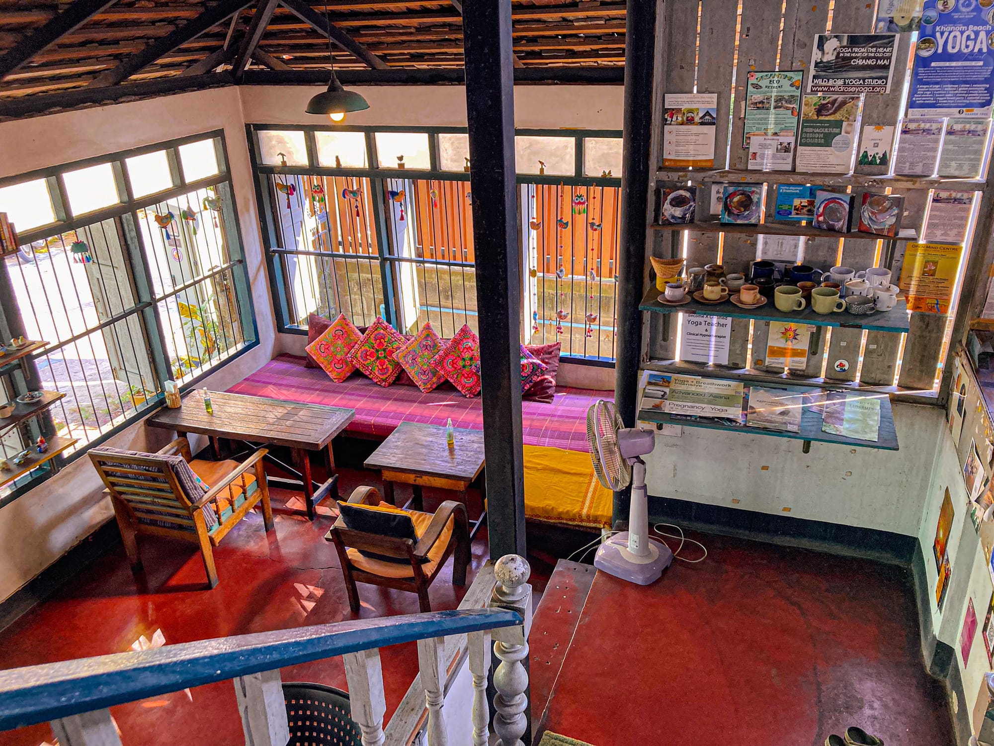 Cozy upstairs seating area at Bird’s Nest Cafe in Chiang Mai with wooden tables, colorful embroidered cushions on a bench by the window, and a small shelf displaying cups and yoga flyers, creating a warm and inviting atmosphere