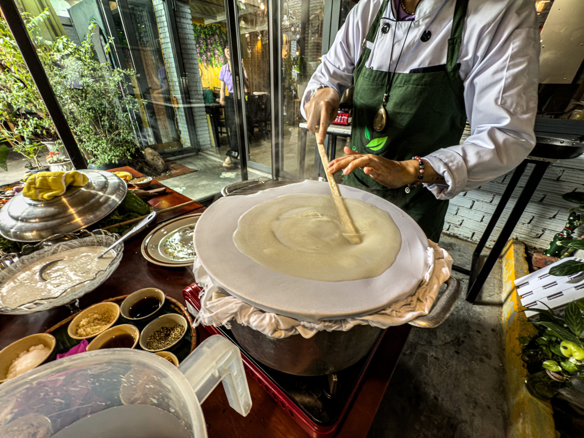 Chef demonstrating how to make fresh rice paper from scratch at Rau Ơi Eatery’s vegan cooking class in Hội An, spreading batter over a steaming cloth with bowls of sauces and ingredients on the side