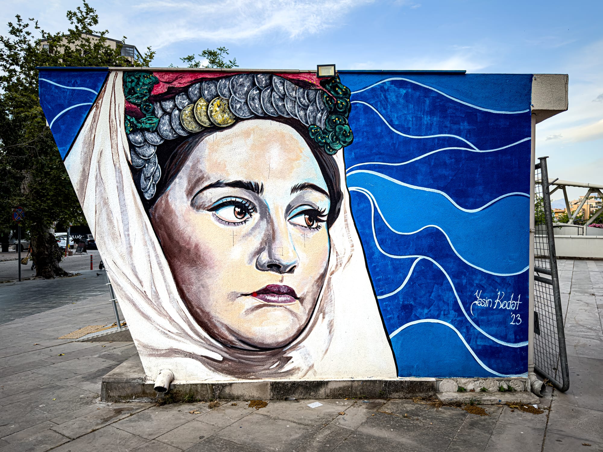 A mural of a woman in traditional headscarf and coin headdress painted on a city building in Antalya, with blue wave patterns behind her