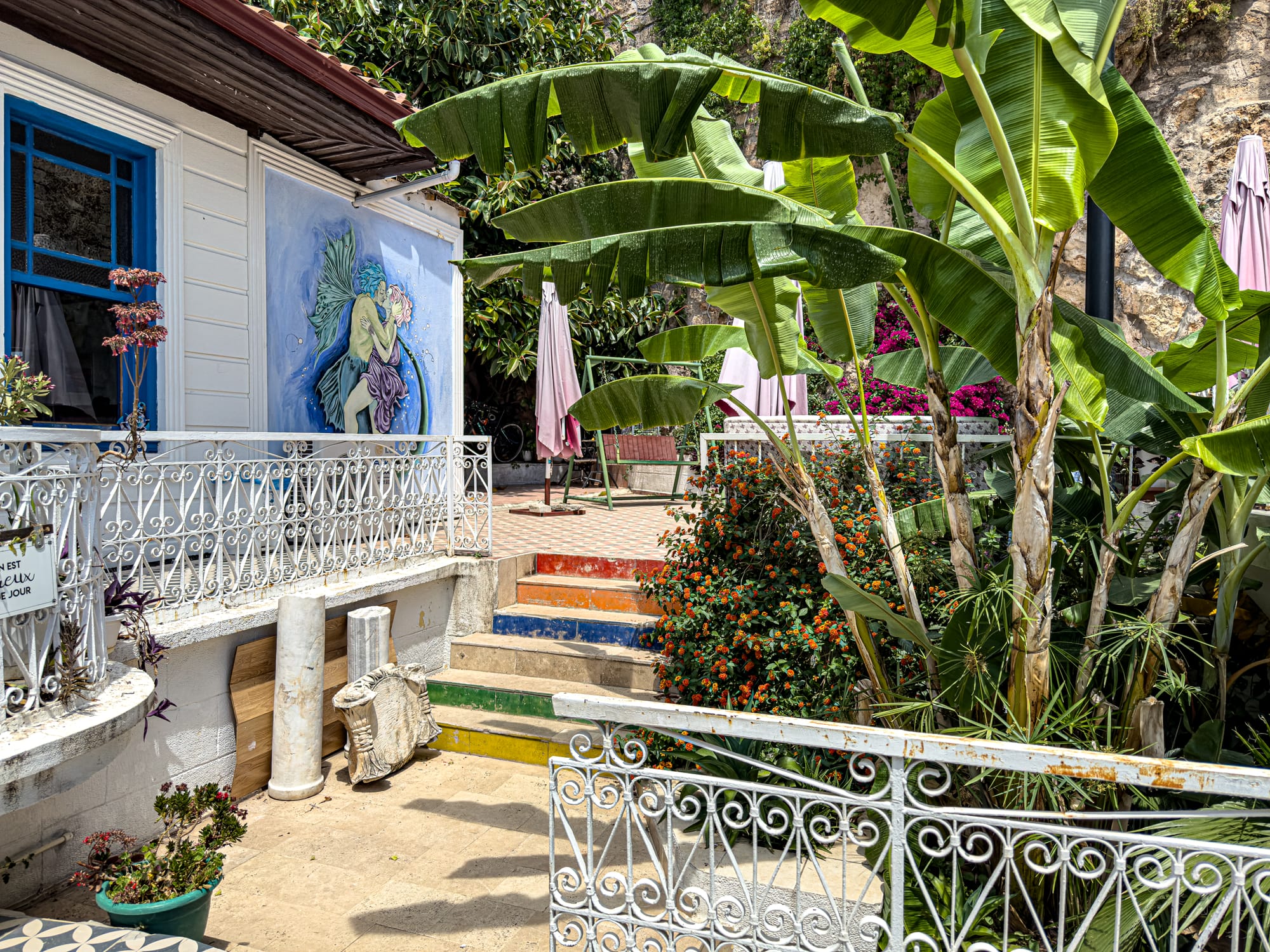 Outdoor terrace at Coworking Antalya featuring lush banana plants, rainbow-painted stairs, and a mural of two figures embracing on the wall