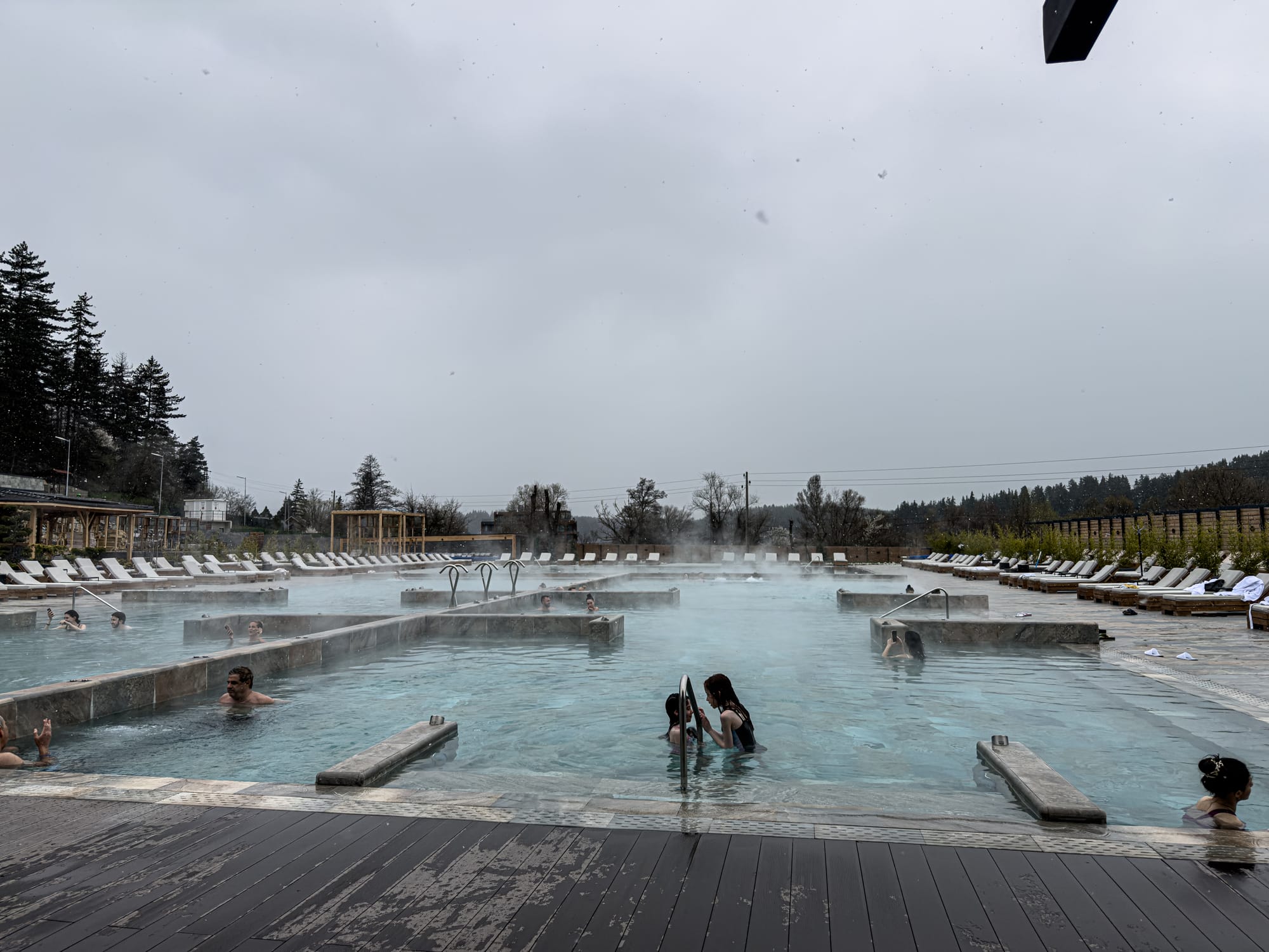 Wide view of steaming mineral pools at Pulse Therme in Banya during winter, with people soaking in the warm water surrounded by rows of empty lounge chairs and a grey, overcast sky