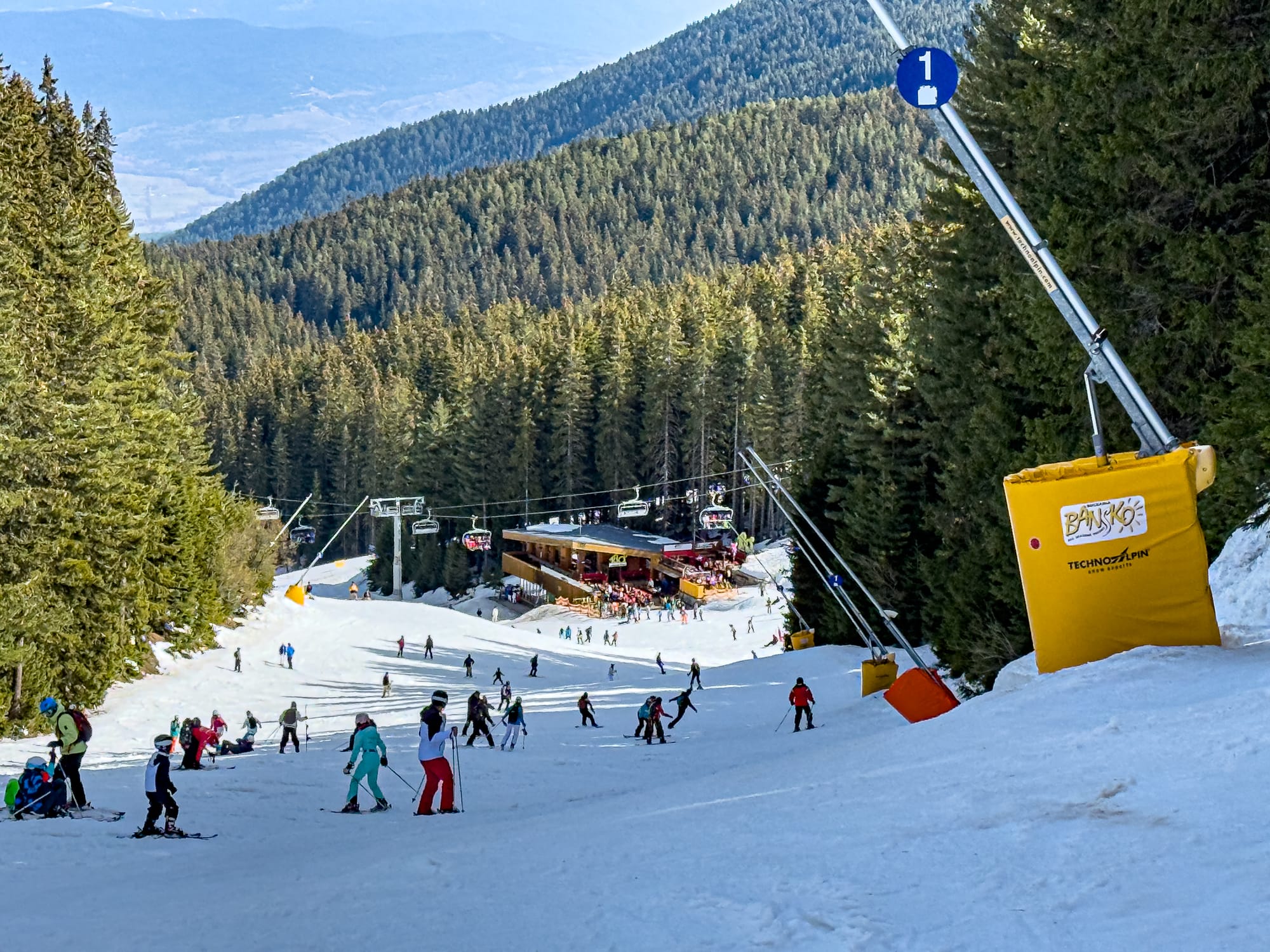 A ski slope in Bansko’s Pirin Mountains filled with skiers and snowboarders, surrounded by dense pine forest, with a mountain hut and ski lifts visible further down the slope