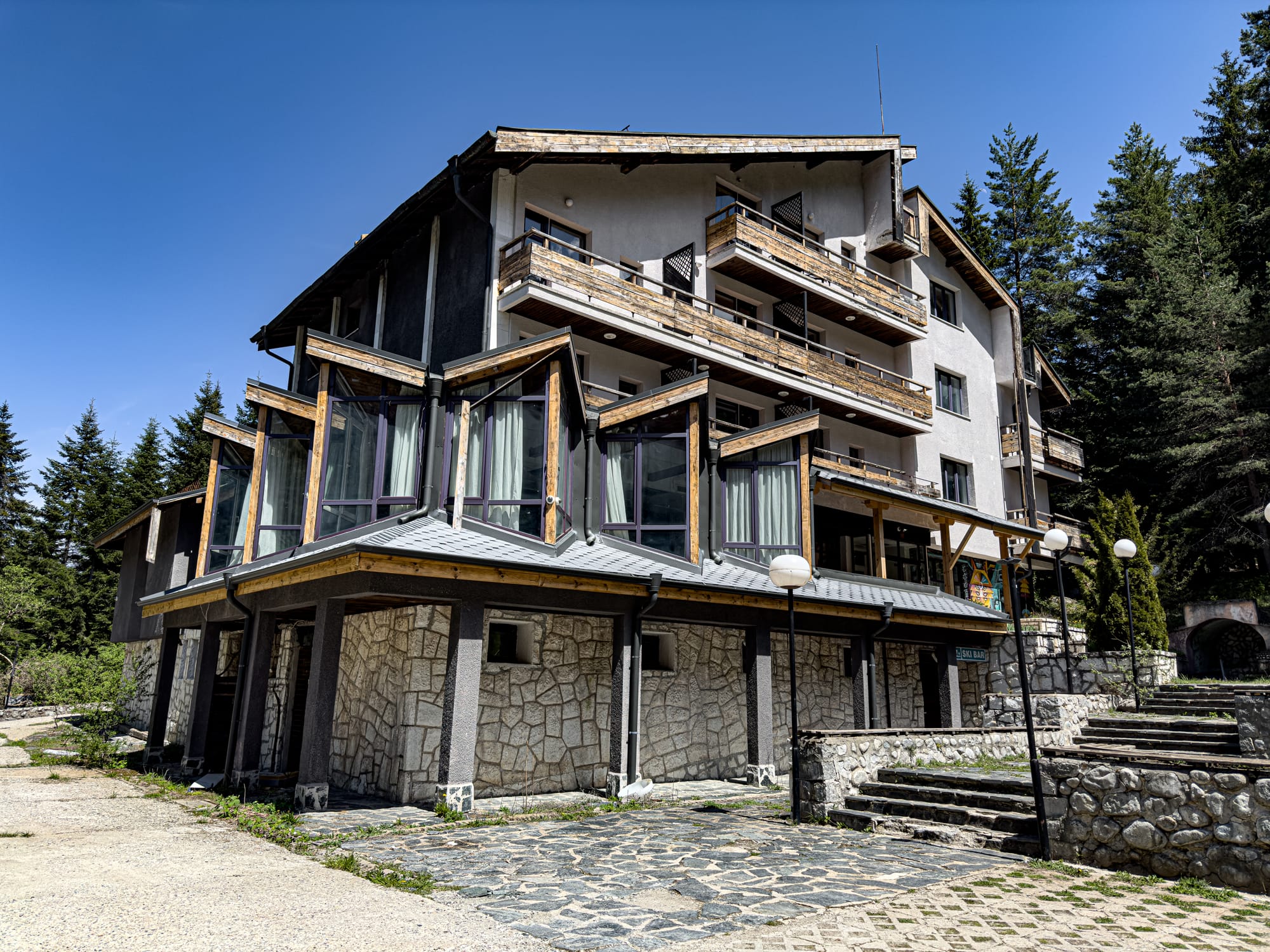 Exterior of the so-called “Secret Hotel” near Bansko, a multi-story stone and wood building with large glass windows, rustic balconies, and forest trees surrounding it under a bright blue sky