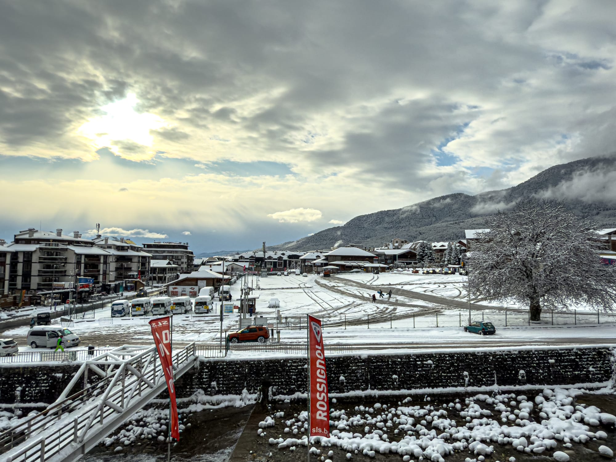 A winter view of Bansko town covered in snow, with modern buildings on the left, a snowy square and parked buses in the center, and the Pirin Mountains rising in the background under dramatic cloudy skies