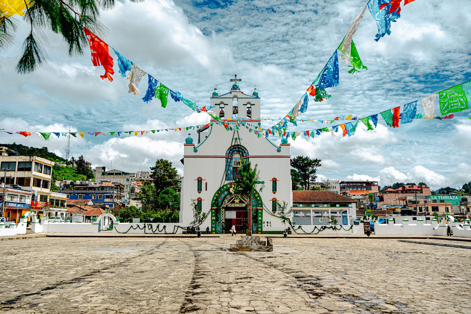 The church of San Juan Bautista in Chamula, Chiapas, decorated with colorful paper flags stretching across the plaza under a bright, cloudy sky, surrounded by the town’s hillside buildings