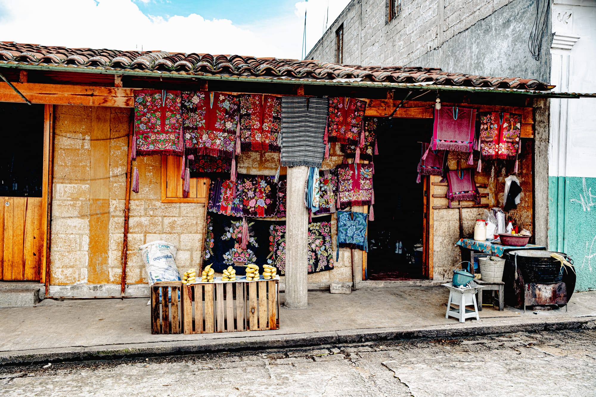 A rustic shop in Zinacantán with handwoven embroidered textiles hanging from the roof, showcasing colorful floral patterns, alongside a small street display of corn and household items
