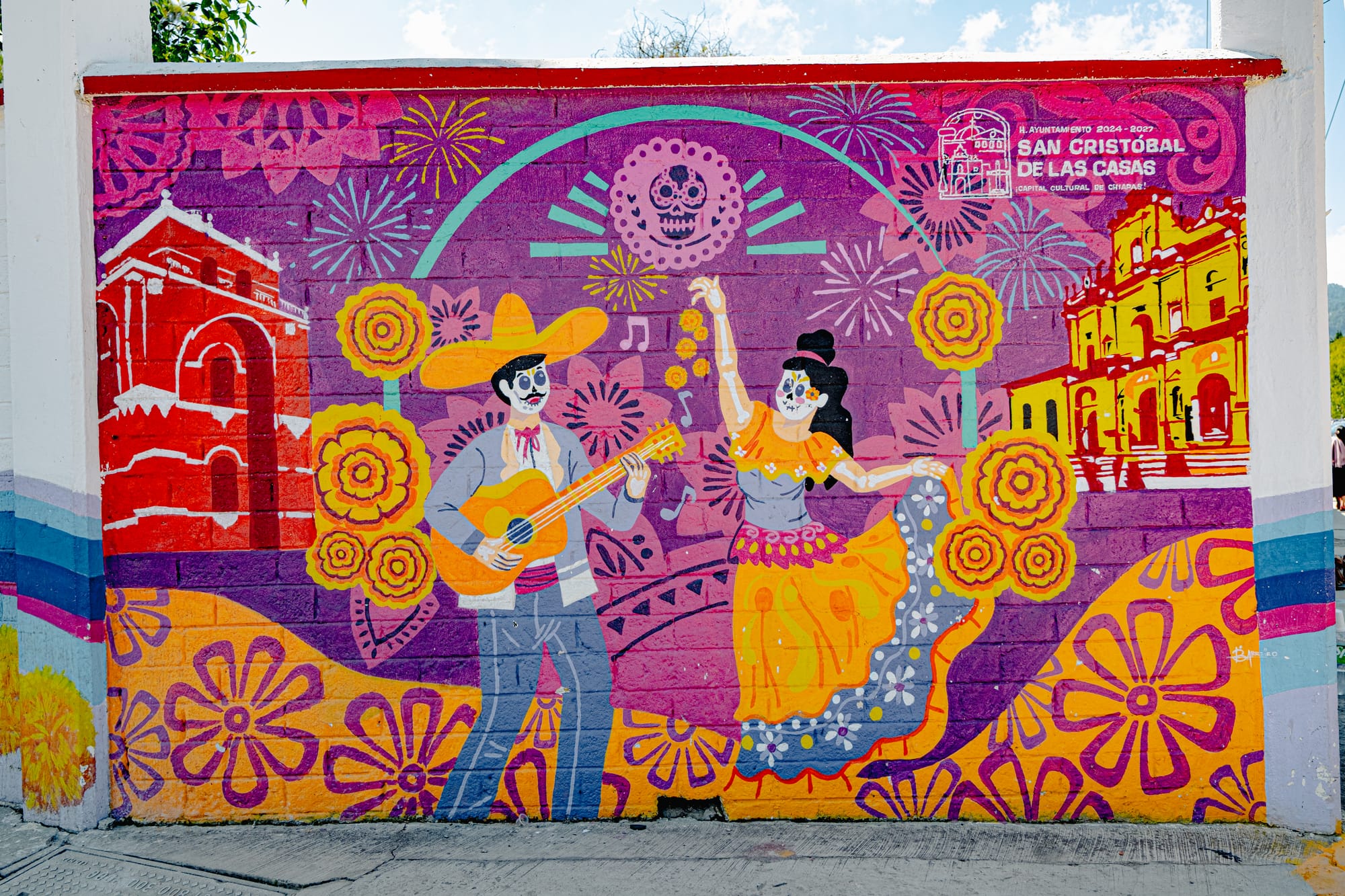 A vibrant mural in San Cristóbal de las Casas depicting skeleton figures of a guitarist and a dancer surrounded by marigolds, fireworks, and local landmarks, celebrating the joy and artistry of Mexico’s Day of the Dead traditions