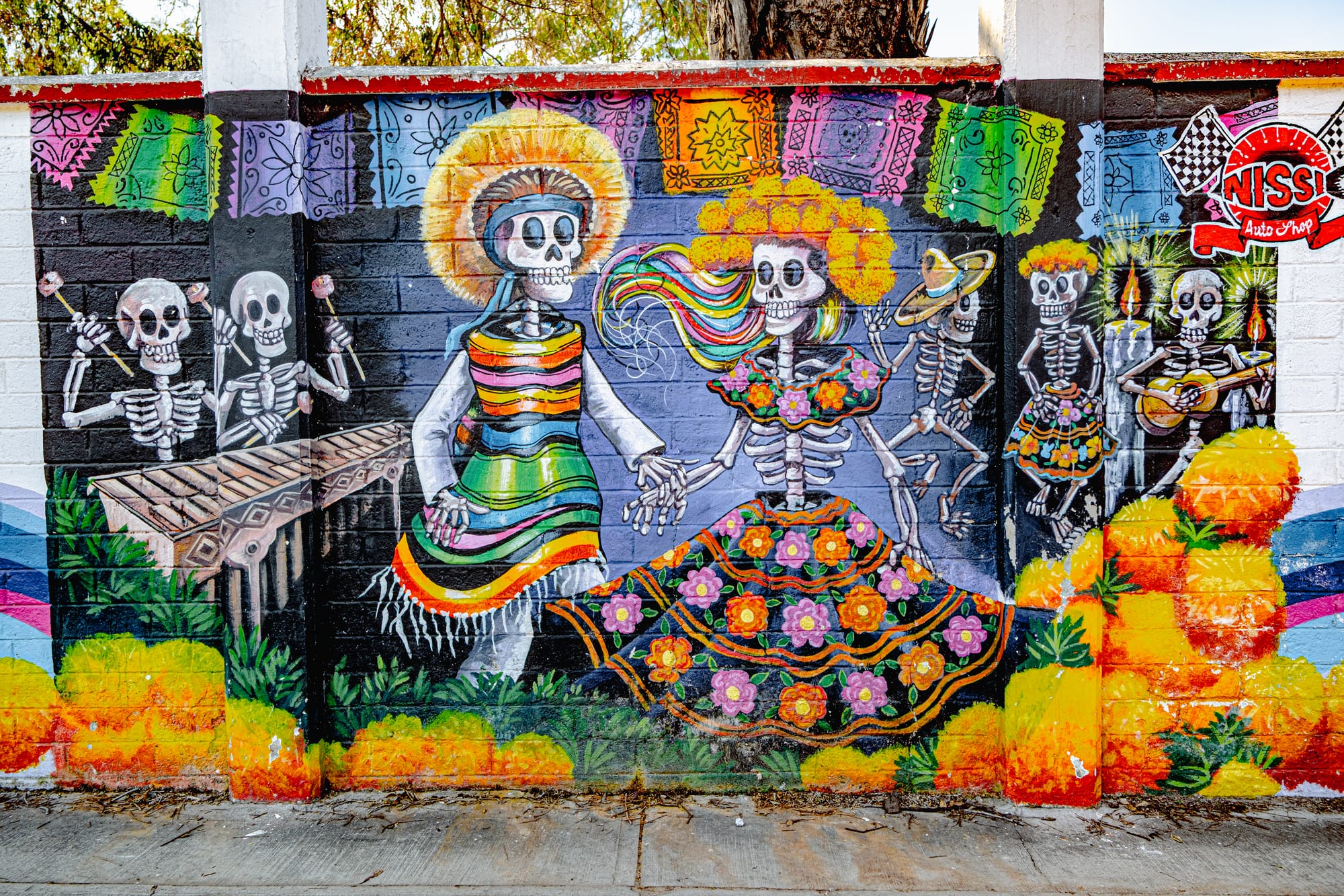 A colorful mural in San Cristóbal de las Casas depicting dancing skeletons in traditional Mexican dress surrounded by marigolds, papel picado, and musicians, celebrating the joyous spirit of Día de los Muertos