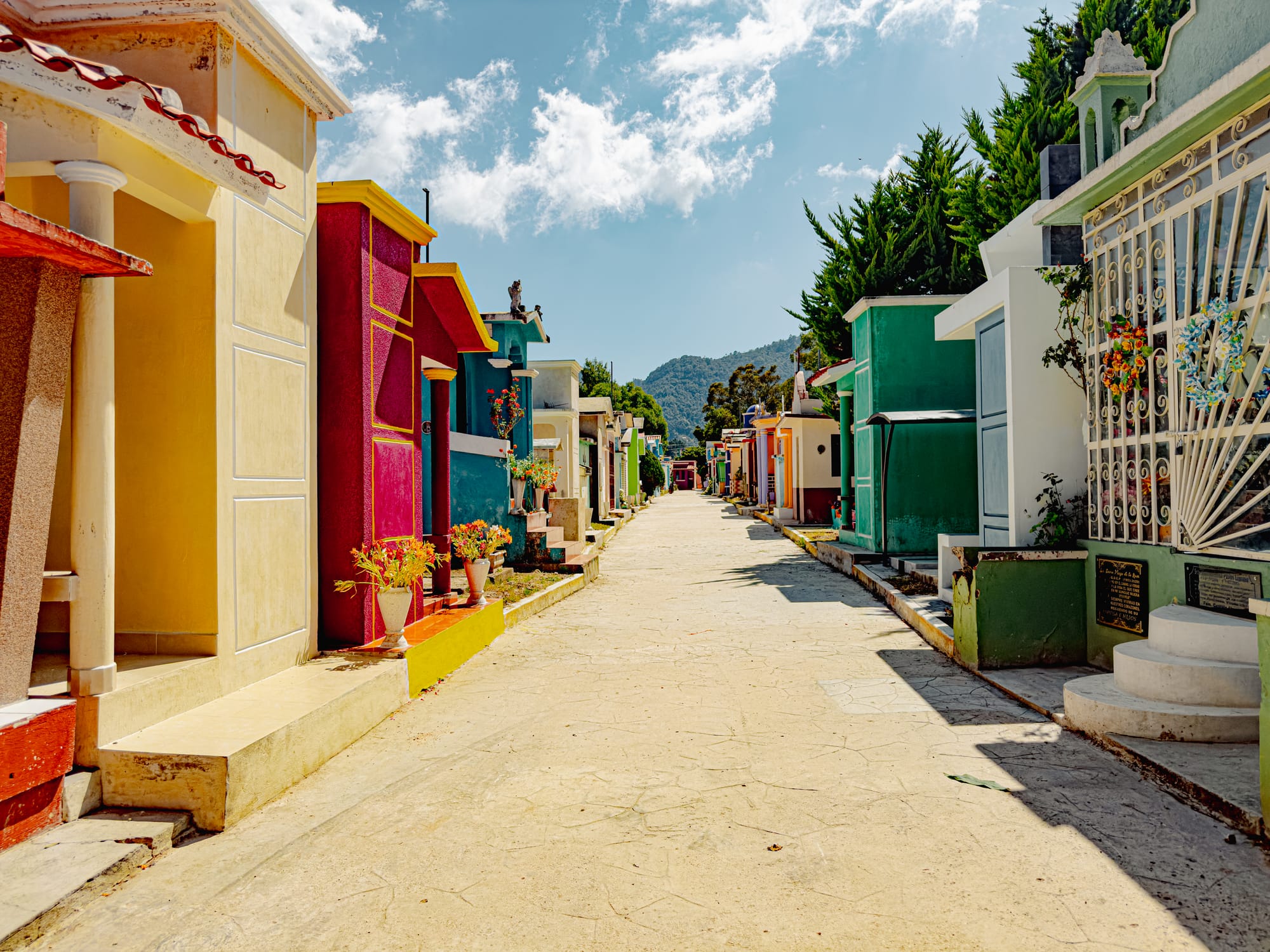 A sunlit walkway through the Municipal Pantheon of San Cristóbal de las Casas, lined with vividly painted tombs in red, yellow, blue, and green, resembling a cheerful street of miniature houses against a backdrop of mountains and bright sky