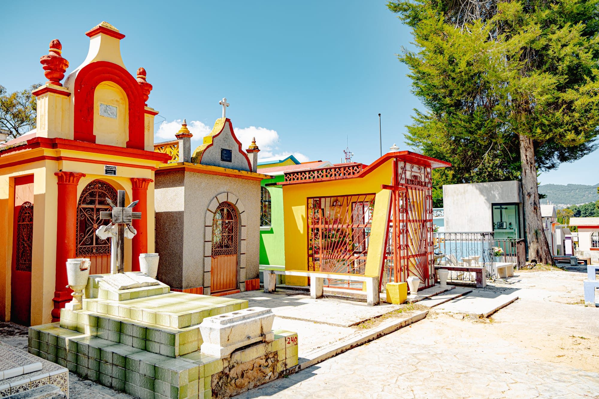 A view of the Municipal Pantheon in San Cristóbal de las Casas showing vibrant yellow, orange, and red tombs with arched doors and crosses, their warm tones illuminated by sunlight beneath a clear Chiapas sky