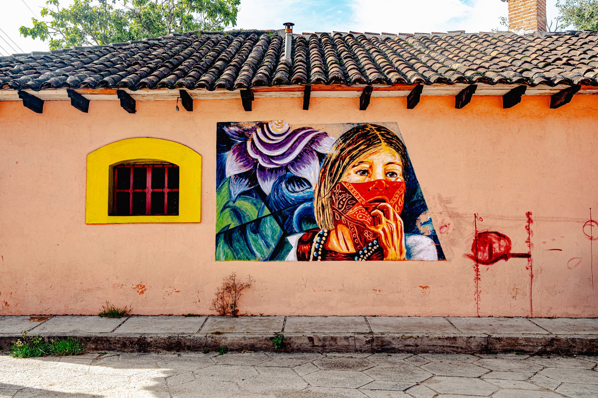 A mural in El Cerrillo, San Cristóbal de las Casas, depicting a young woman wearing a red bandana over her face against a backdrop of abstract flowers and leaves, symbolizing Indigenous resistance, dignity, and the enduring presence of Zapatista identity in Chiapas