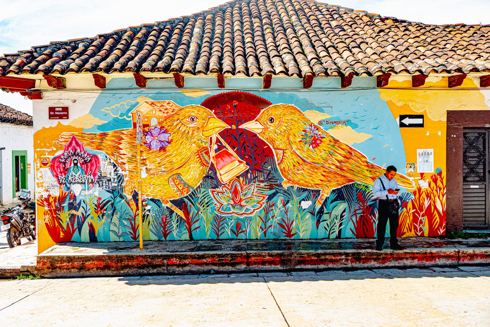 A colorful mural in El Cerrillo, San Cristóbal de las Casas, showing two large yellow birds exchanging a red bell amid flowers and plants on a tiled-roof corner building, with a man standing in front checking his phone