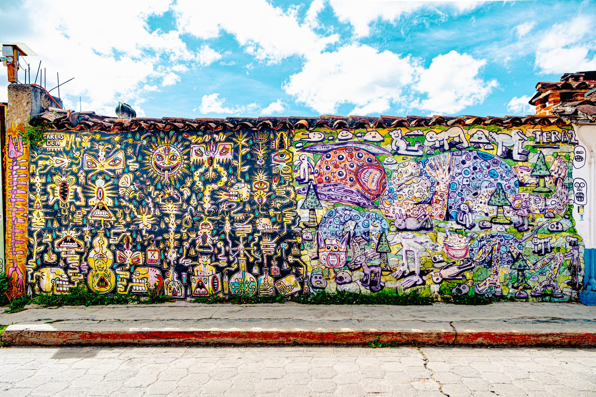 A large mural in El Cerrillo, San Cristóbal de las Casas, by artist Carlos Cea, featuring intricate symbols, faces, and surreal creatures in vivid yellows, purples, and greens, blending myth, dream imagery, and local cosmology into a psychedelic tapestry of Chiapas street art