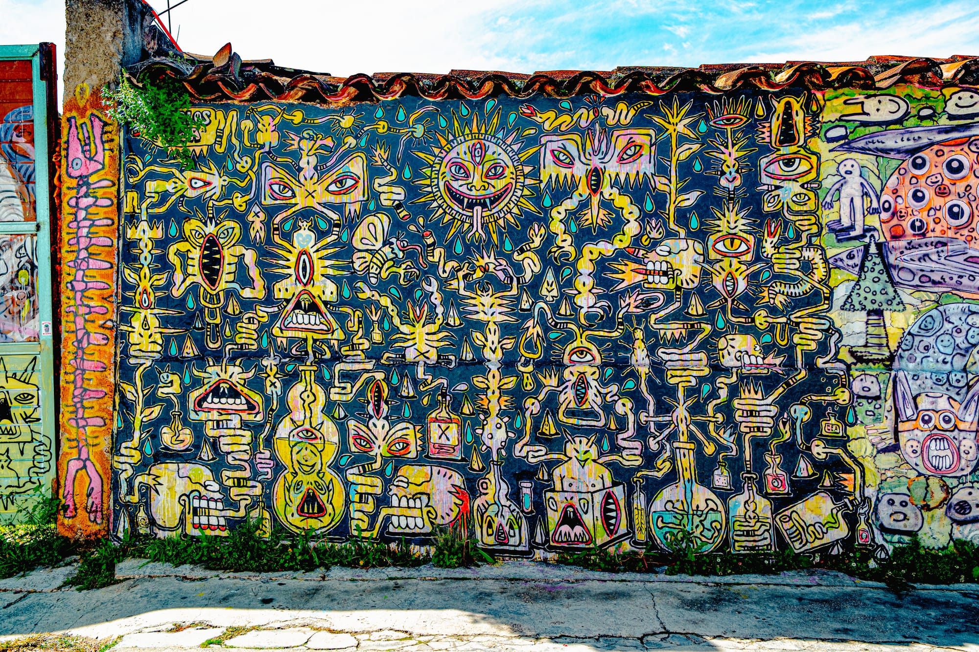A detailed mural by artist Carlos Cea in El Cerrillo, San Cristóbal de las Casas, filled with intricate yellow and pink symbols, eyes, suns, and abstract figures on a dark blue background, evoking cosmology, mysticism, and the vibrant visual language of Chiapas street art