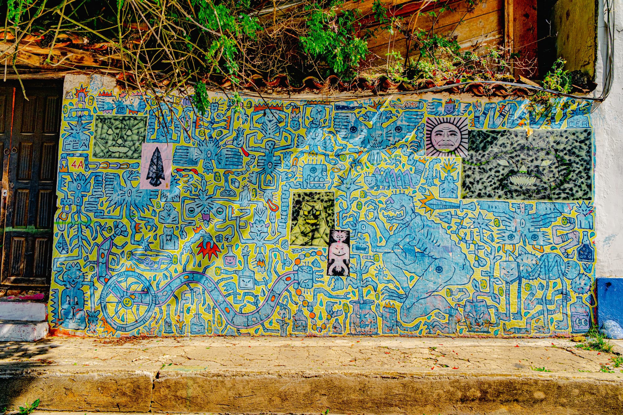 A mural by Carlos Cea in El Cerrillo, San Cristóbal de las Casas, filled with intricate blue and yellow symbols, mythic figures, and surreal shapes framed by leafy branches, blending pre-Hispanic motifs with dreamlike contemporary expression