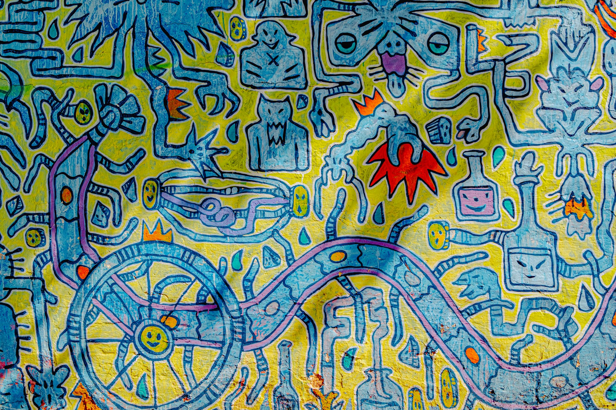 Close-up of a Carlos Cea mural in El Cerrillo, San Cristóbal de las Casas, showing blue and purple abstract figures, tubes, bottles, and a wheel on a yellow background, forming a dense, surreal network of interconnected symbols and playful shapes